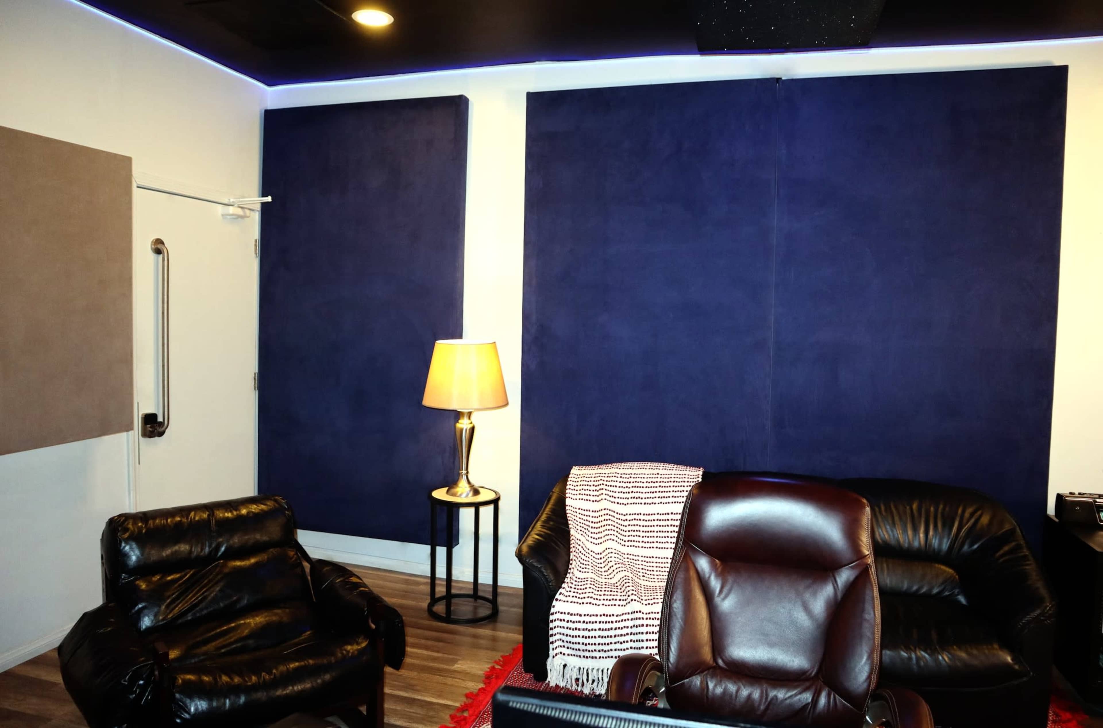 The image shows a soundproof room with blue acoustic panels on the walls, a standing lamp, and leather chairs arranged around a small table.