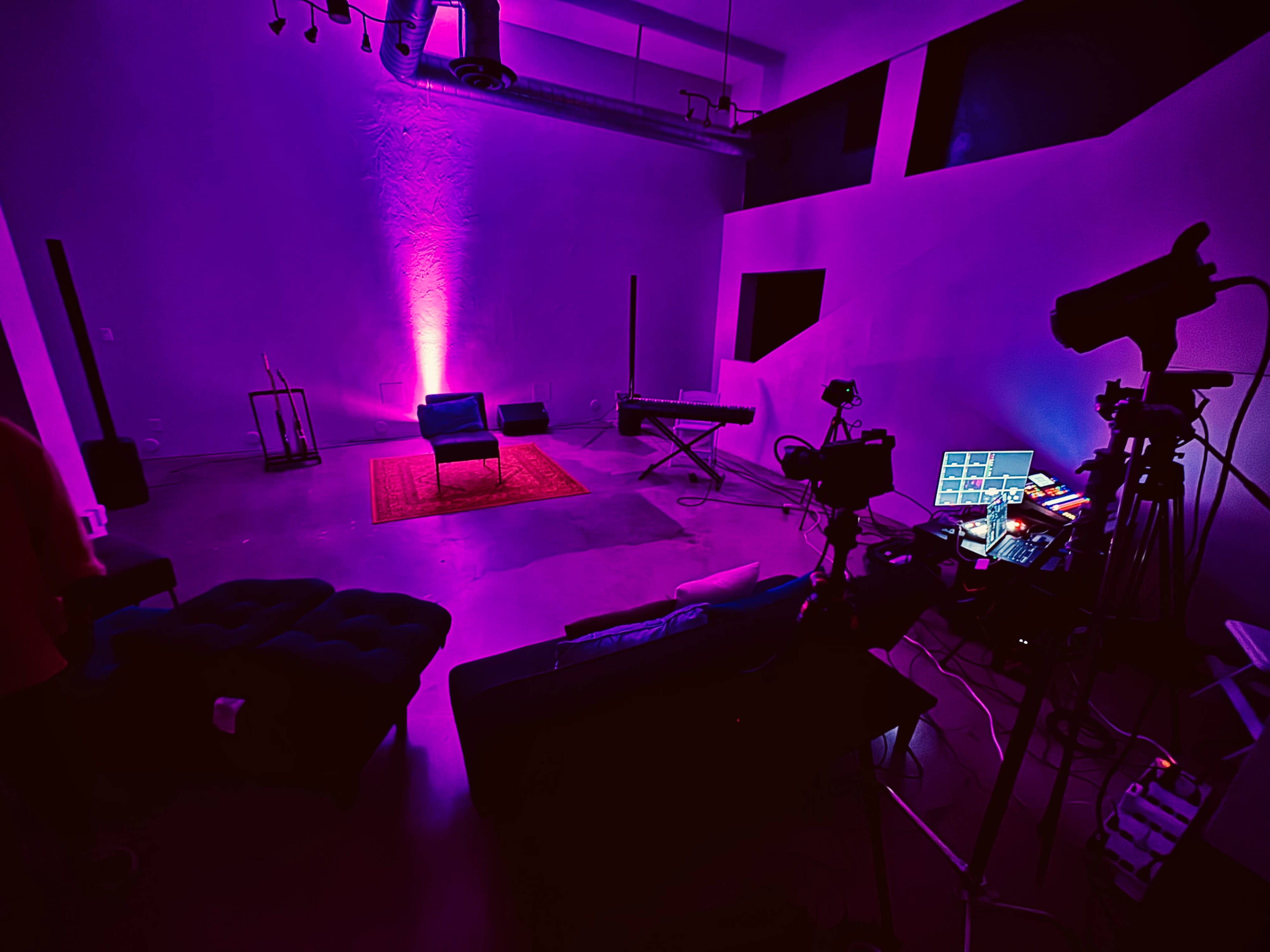The image shows a spacious room with purple lighting, featuring two blue chairs, a keyboard, a rug, and various cameras and equipment set up for recording.
