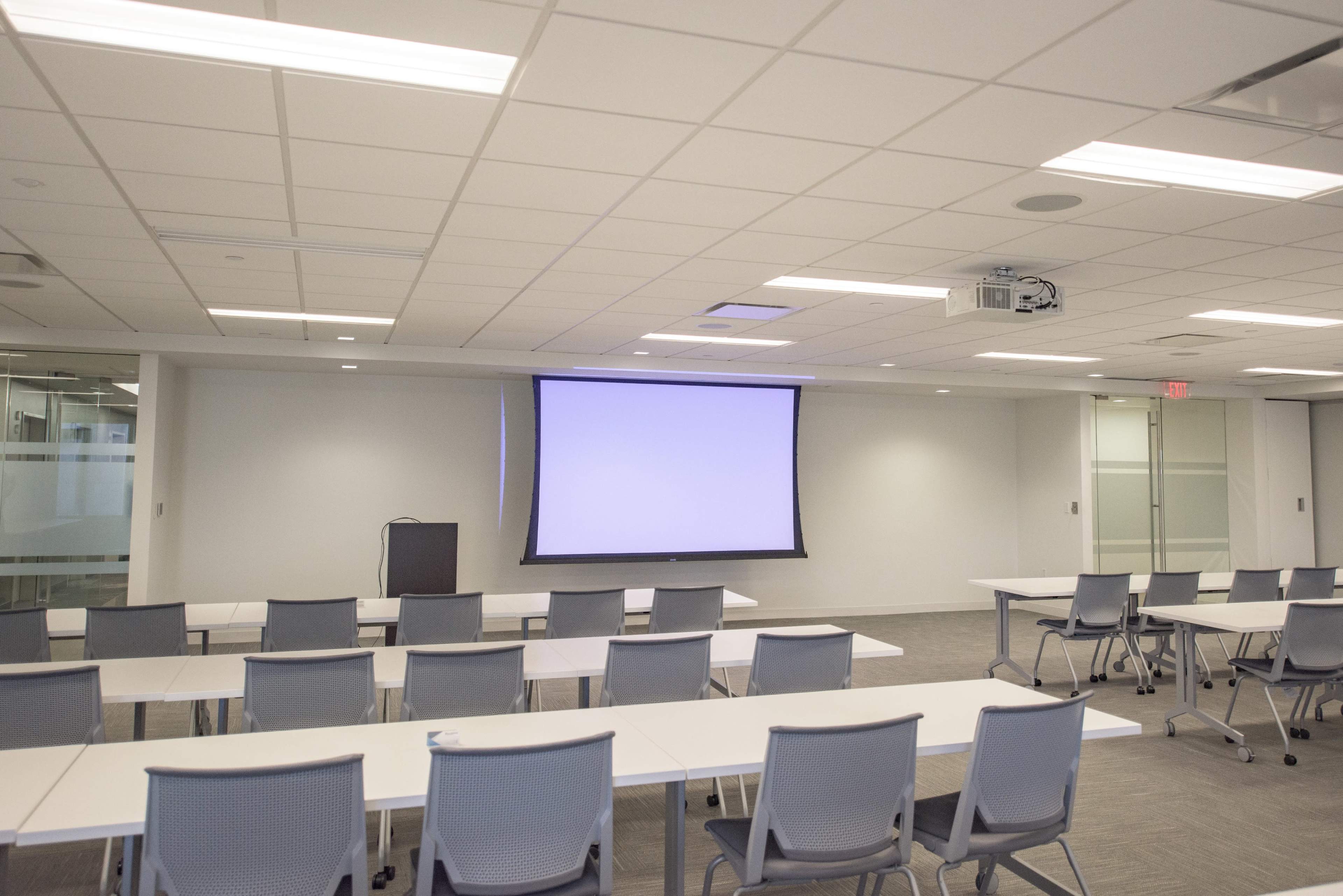 A spacious conference room features several rows of tables and chairs arranged neatly, with a large screen at the front and a projector mounted on the ceiling.