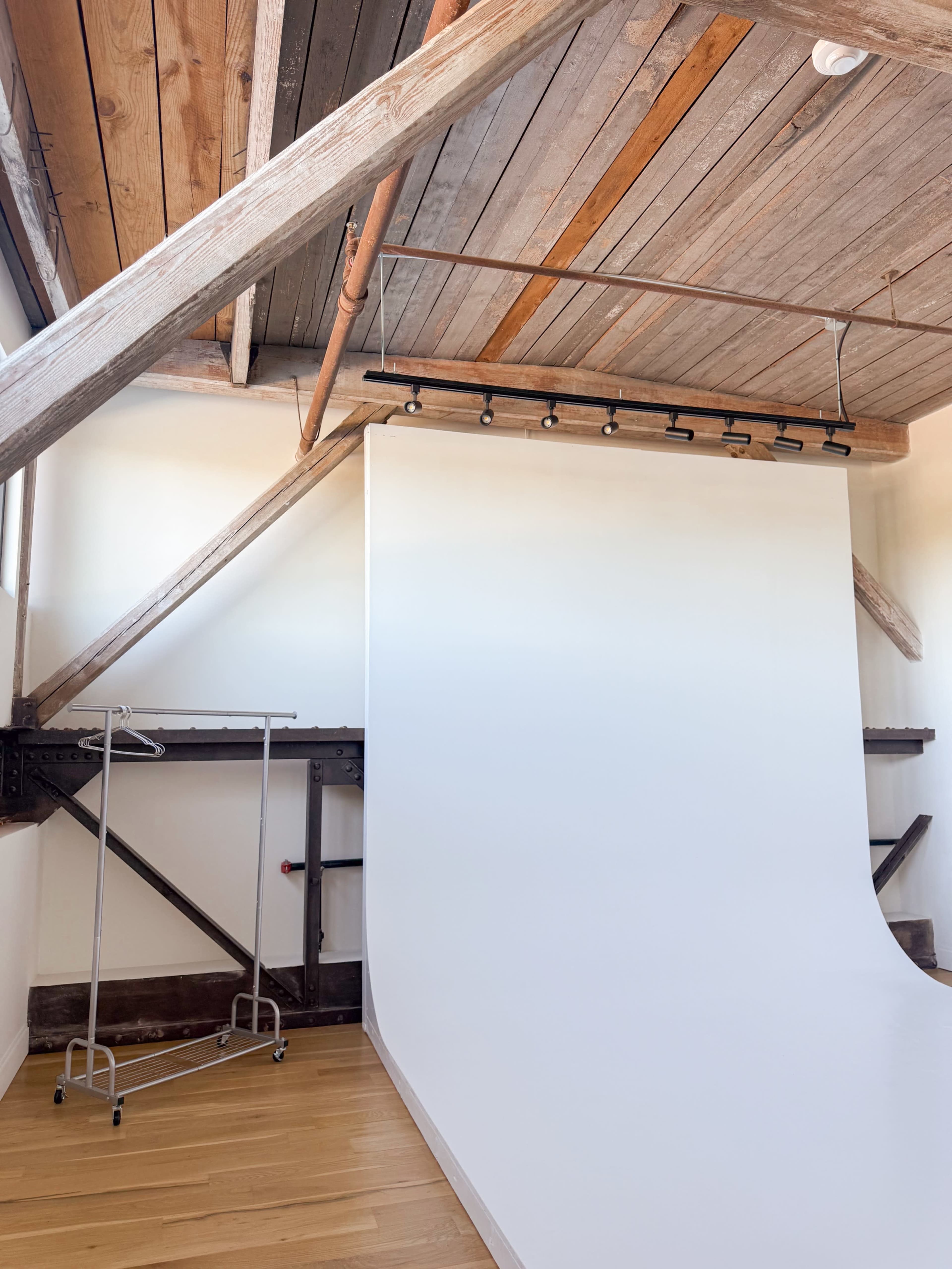 The image shows a studio space featuring a white backdrop, wooden beams, and a metal clothing rack.