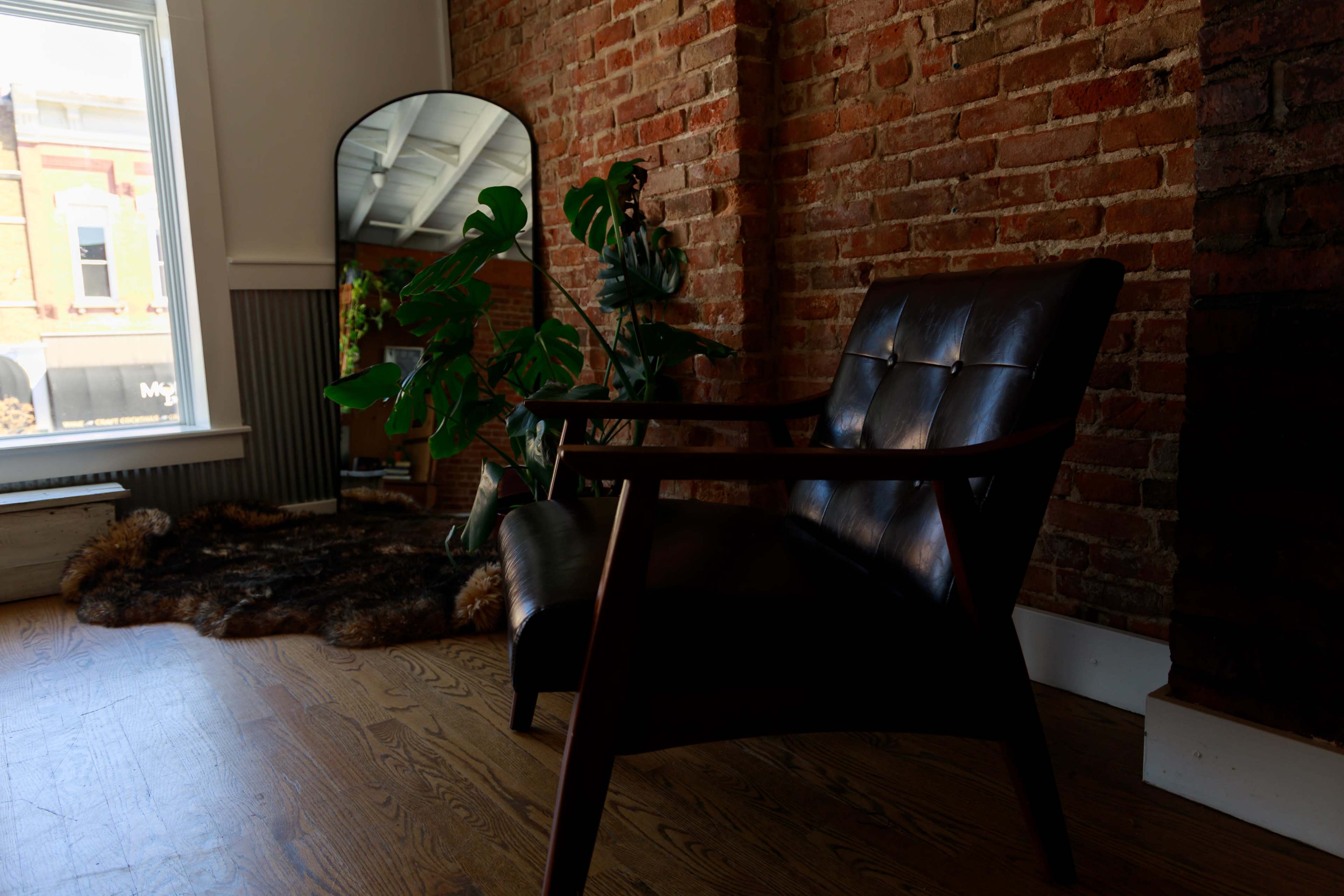 A leather chair is positioned beside a plant and a mirror against a brick wall in a room with wooden flooring.