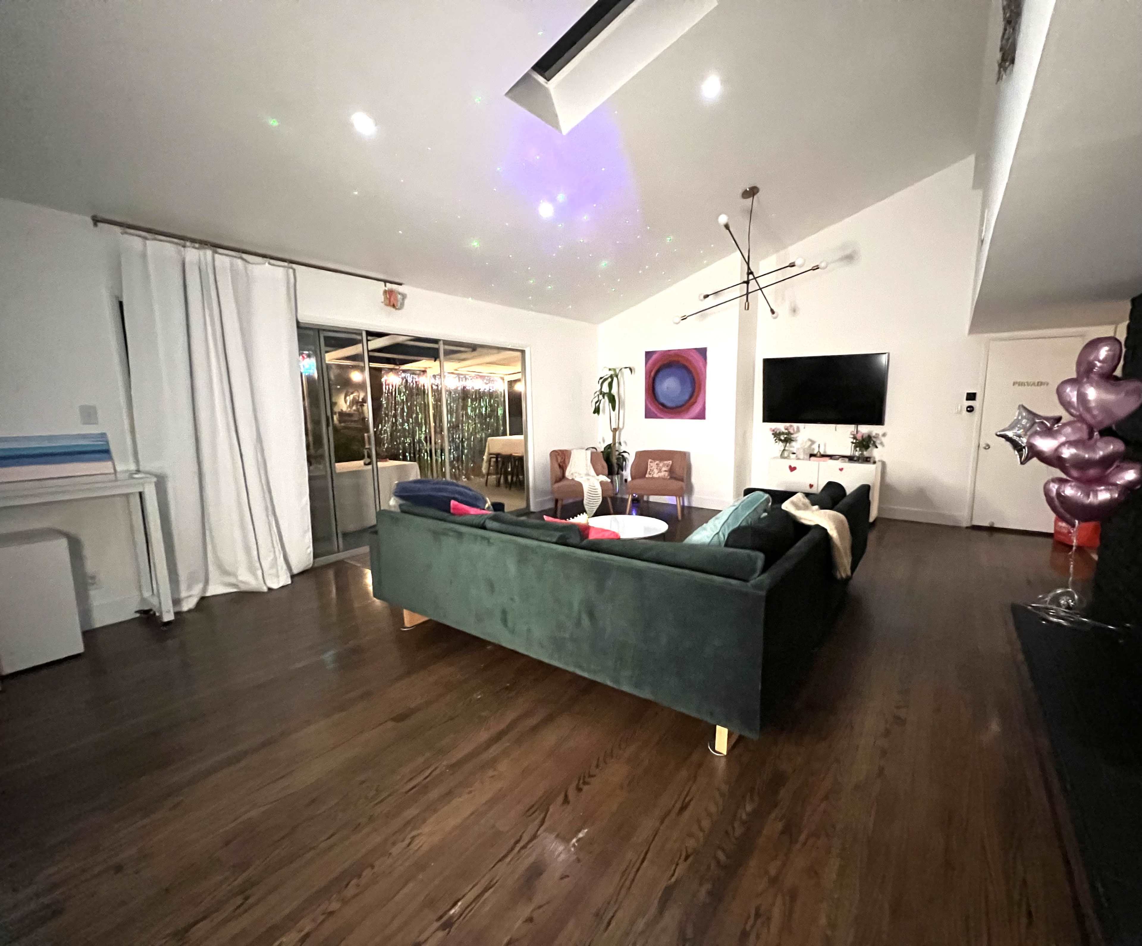 A spacious living room features a green sofa, a large television, and a backdrop of colorful lights reflecting on the ceiling.
