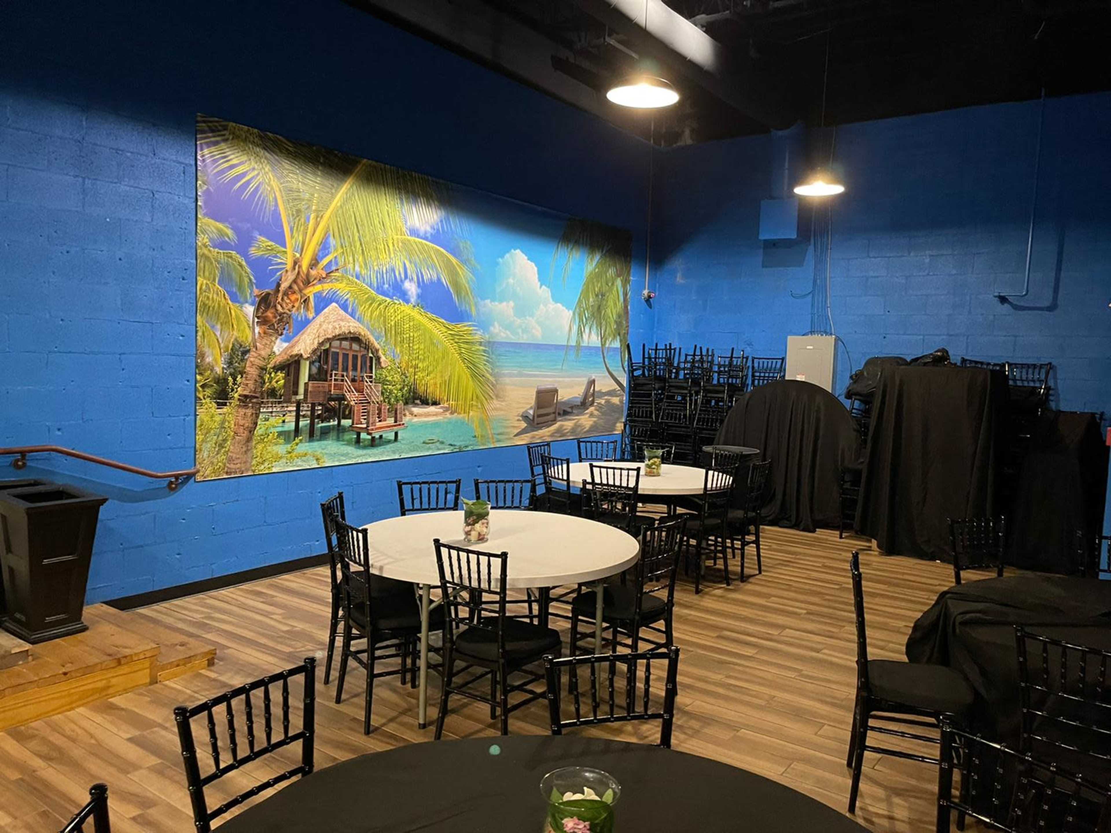 An event space with circular tables and black chairs, featuring a large tropical mural on a blue-painted wall.