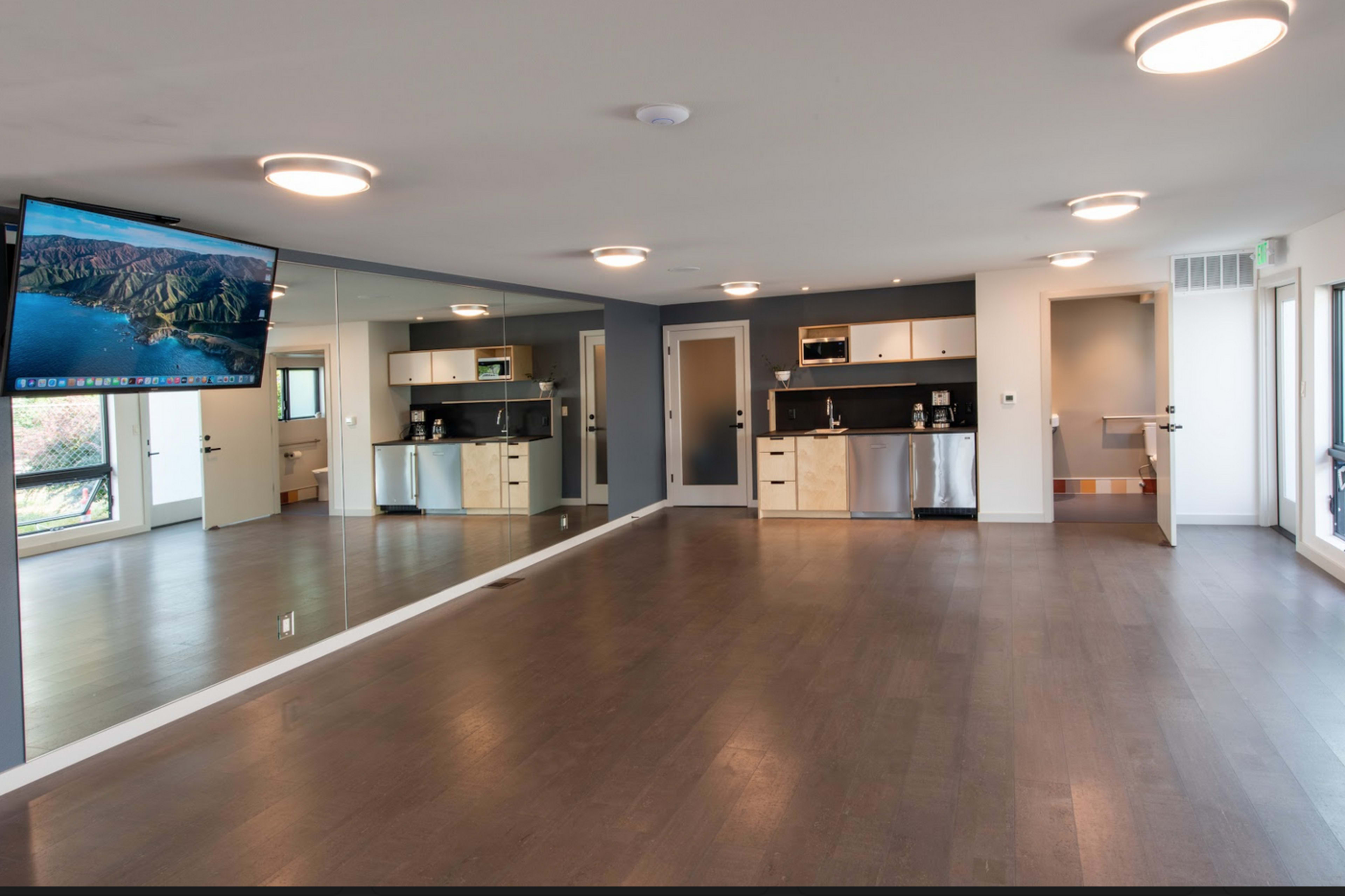 A spacious room with a kitchen area featuring modern appliances, hardwood flooring, and a large mirror reflecting the space.