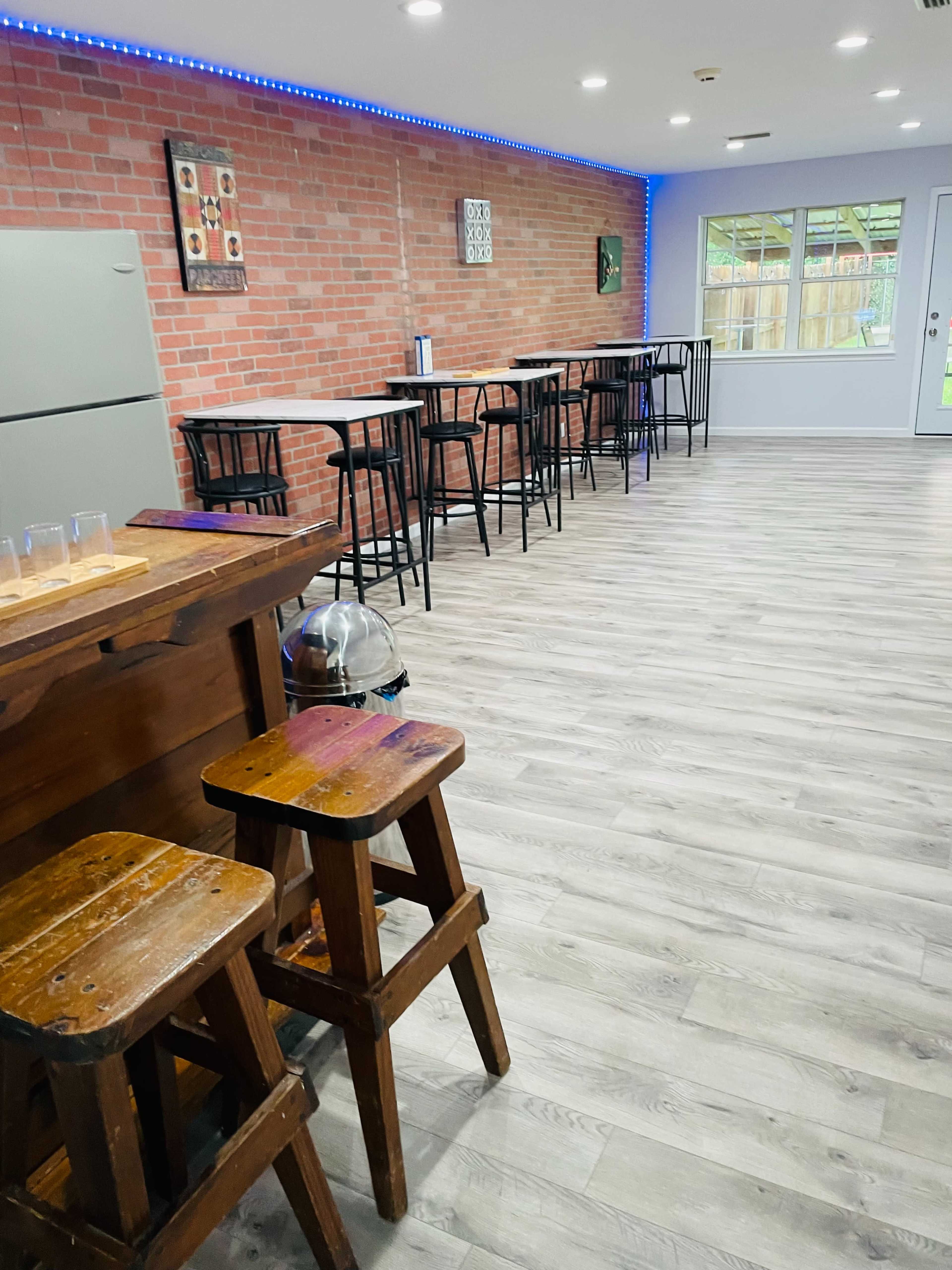 A room featuring a bar area with wooden stools, a refrigerator against a brick wall, and several high-top tables with chairs arranged along the opposite wall.