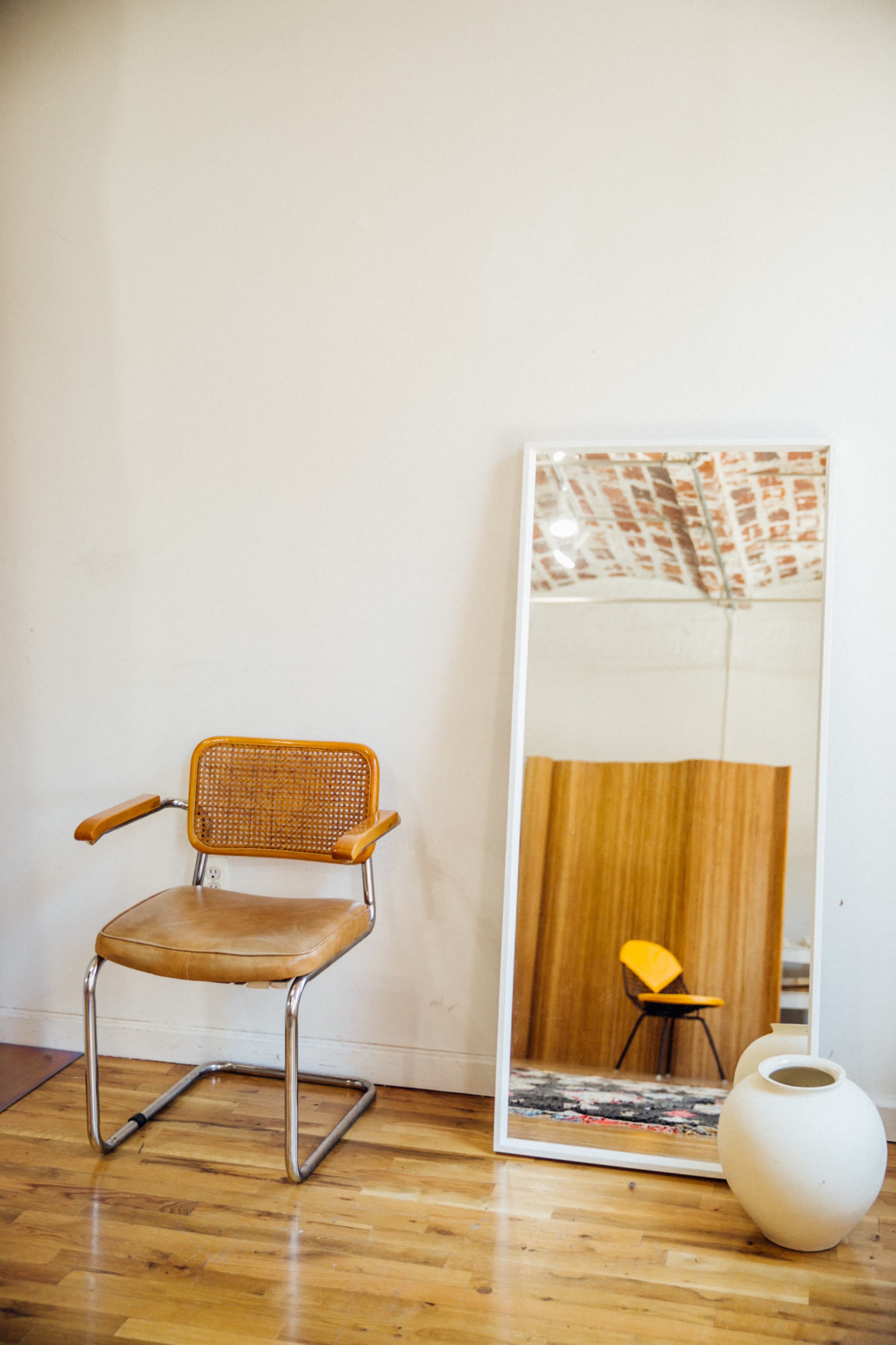 A chair with a woven backrest stands beside a large mirror reflecting a wooden panel and another chair in a minimalist interior space.