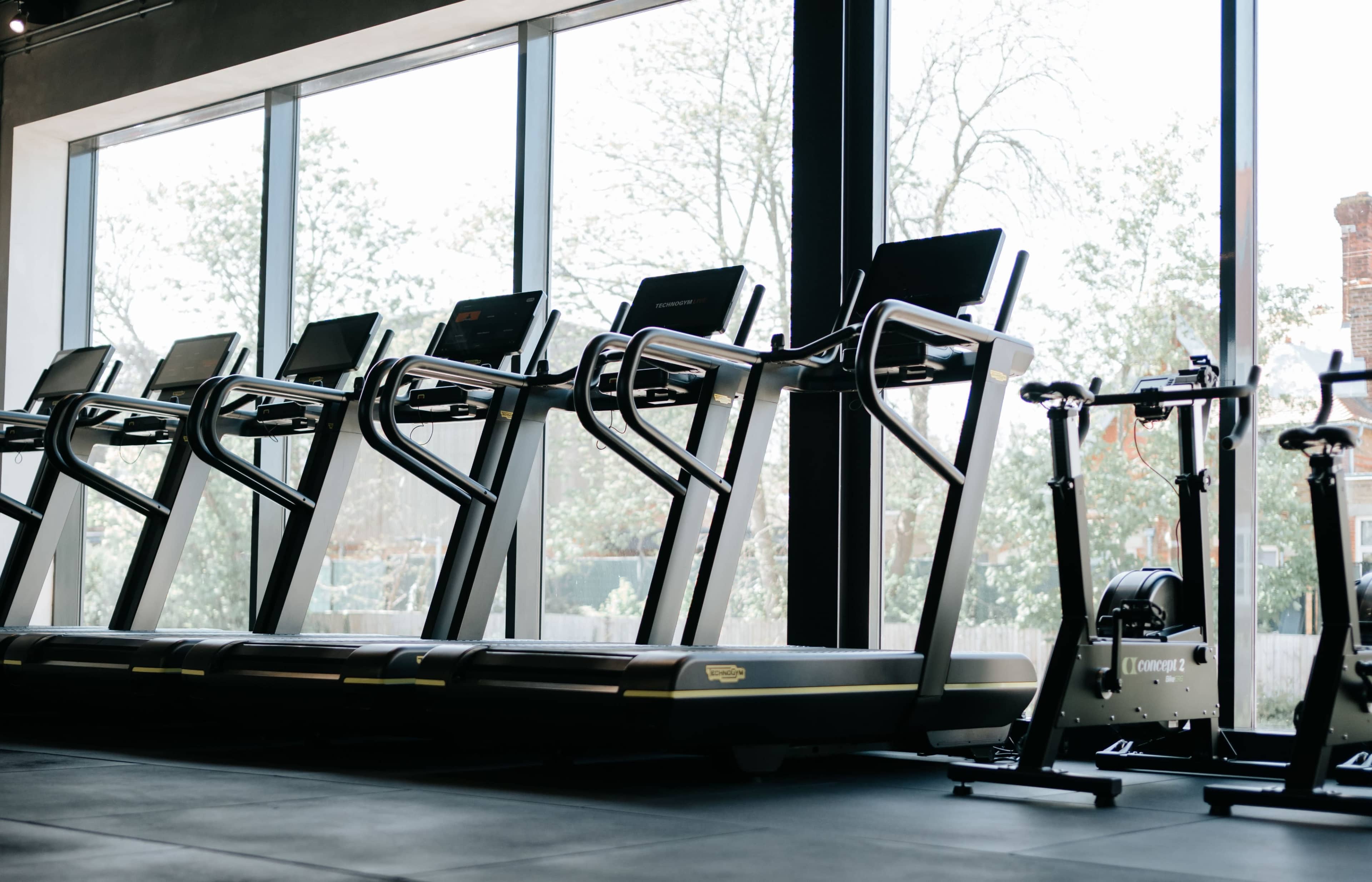 The image shows a line of treadmills positioned in front of large windows in a gym.