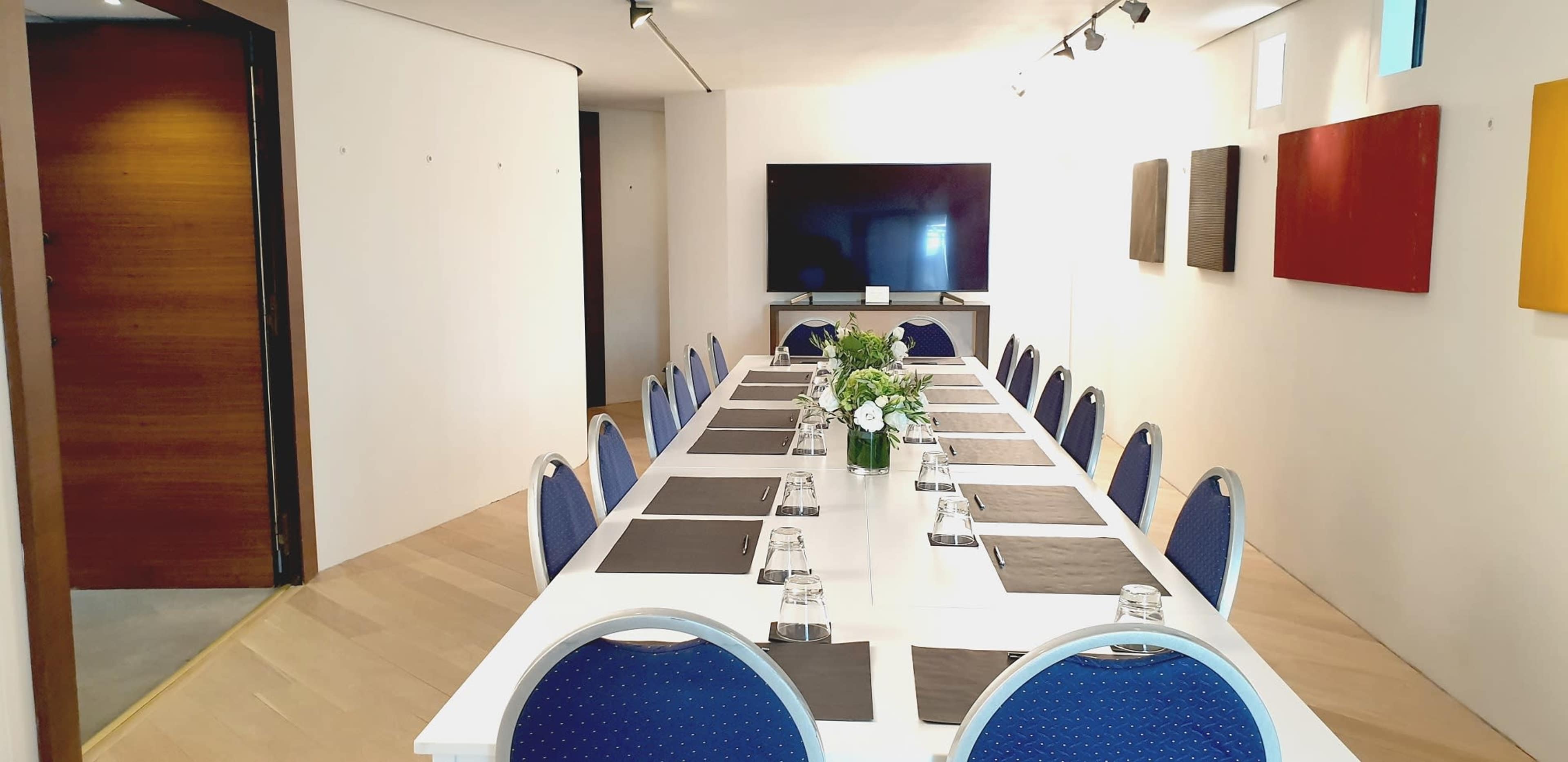 A conference room features a long white table surrounded by blue chairs, with a television screen at one end and framed artwork on the walls.