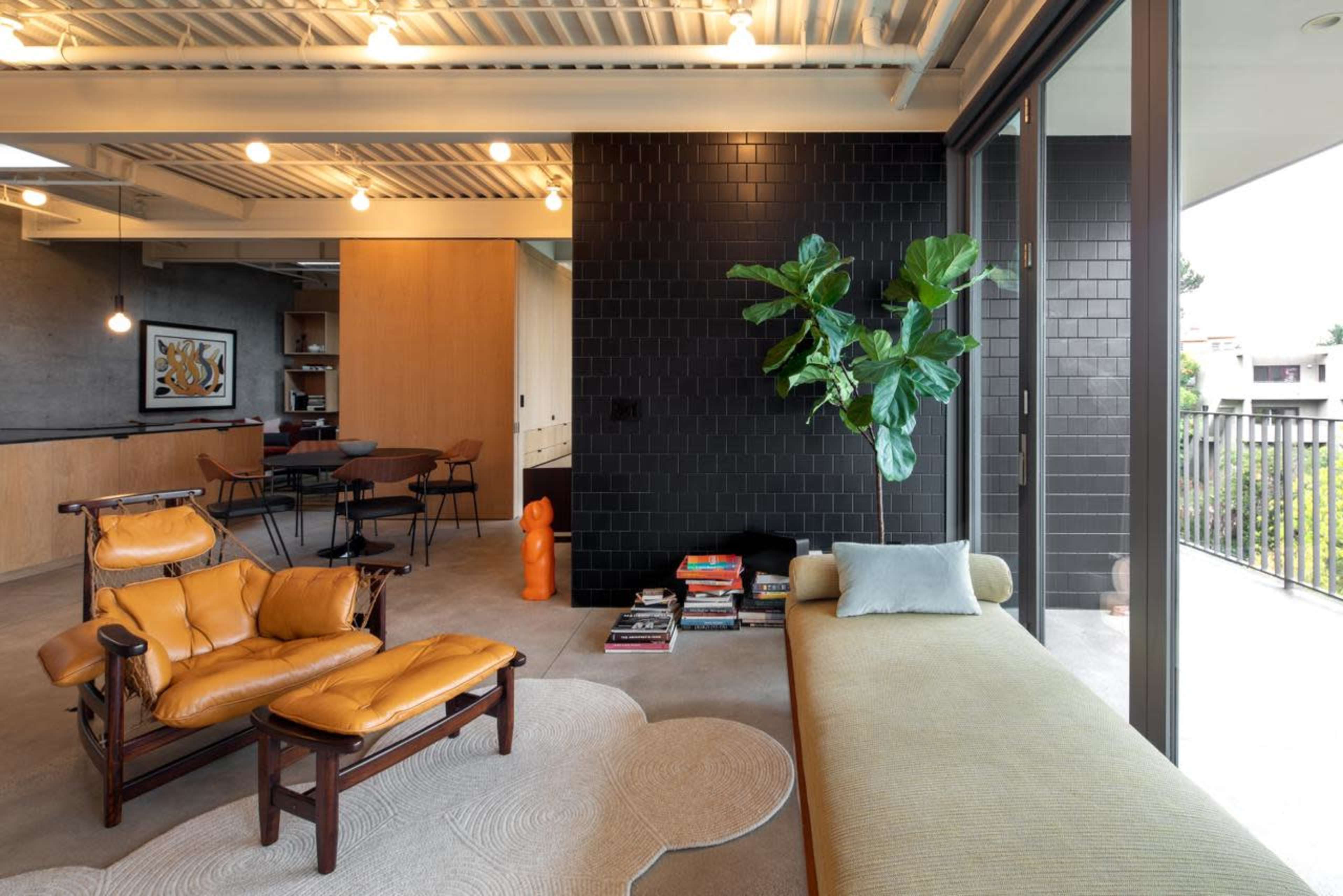 The interior of a modern living room features a yellow leather chair and a light green couch, complemented by a dark tiled wall and large windows that open to a balcony.