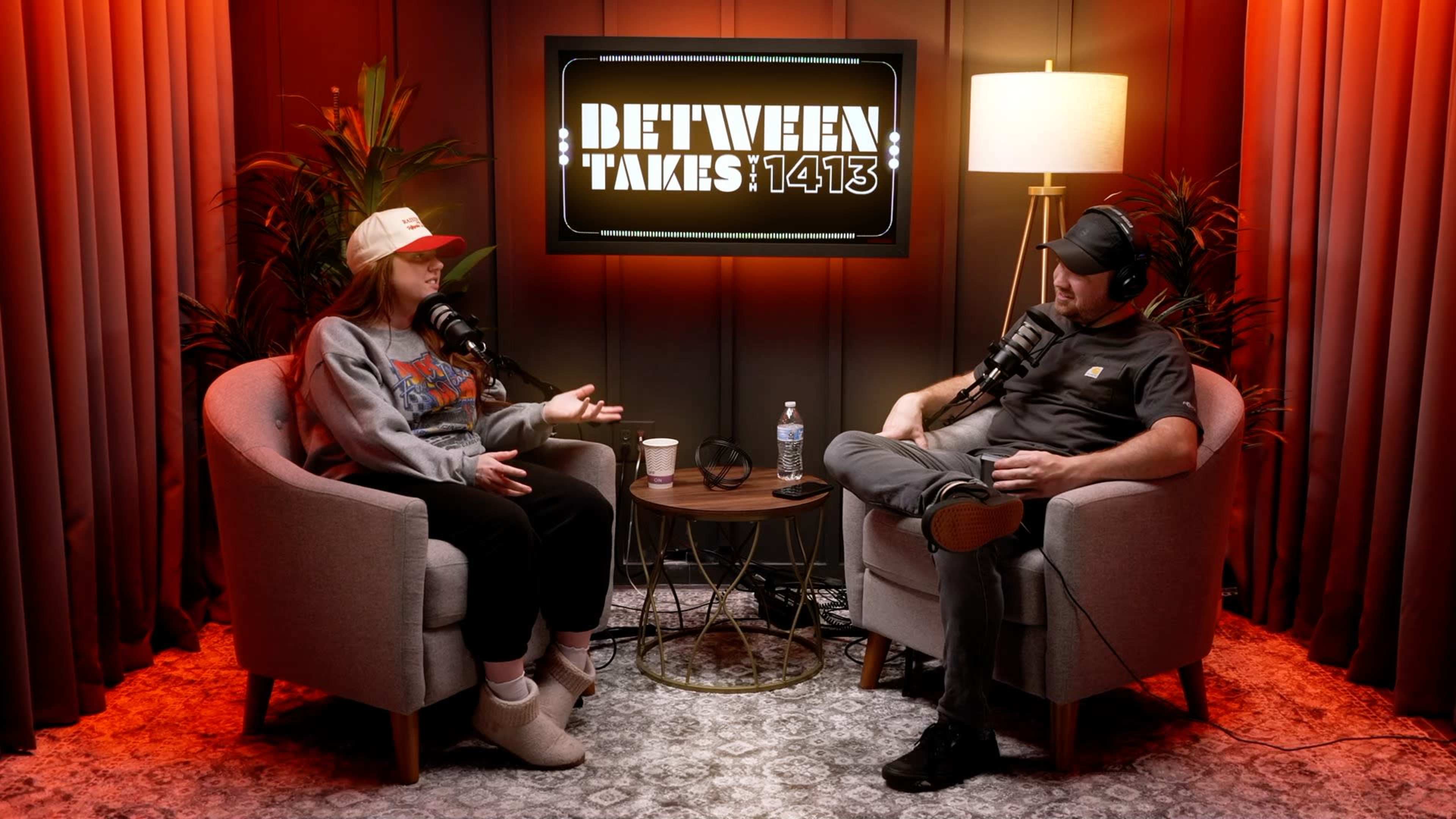 A woman and a man are seated in modern armchairs in a podcast studio, discussing topics while microphones are positioned in front of them.