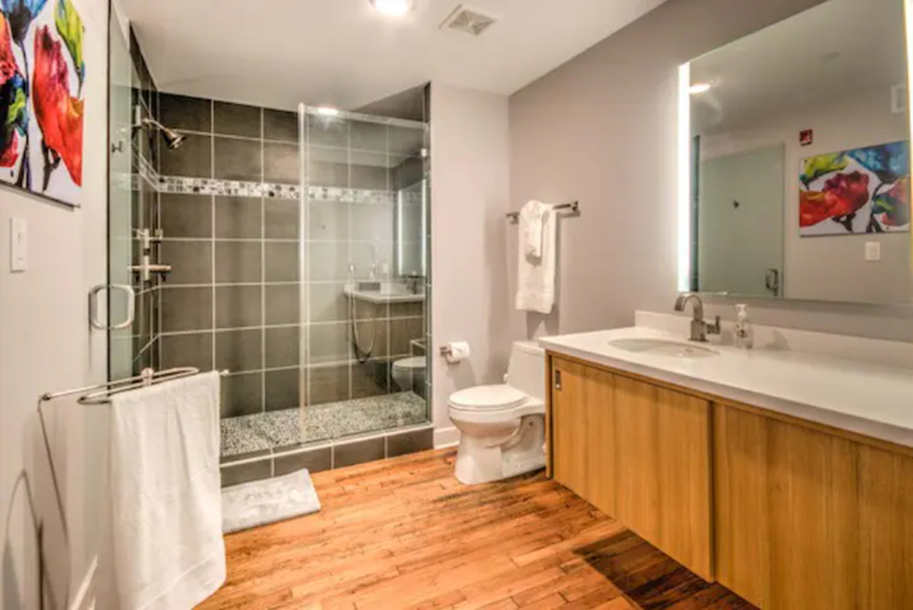 A modern bathroom featuring a glass shower, wooden flooring, a white countertop with a sink, and bright artwork on the walls.