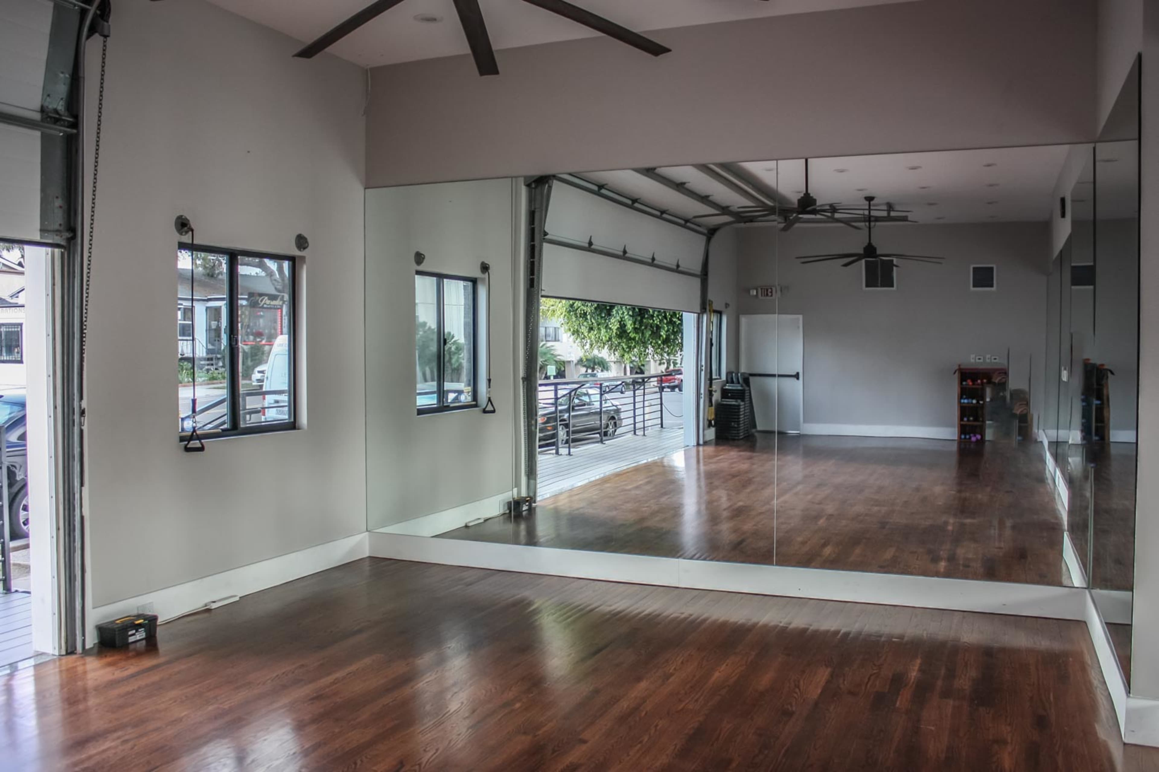 A spacious room with wooden floors, large mirrors on one wall, and a garage-style door partially open, allowing natural light to enter.