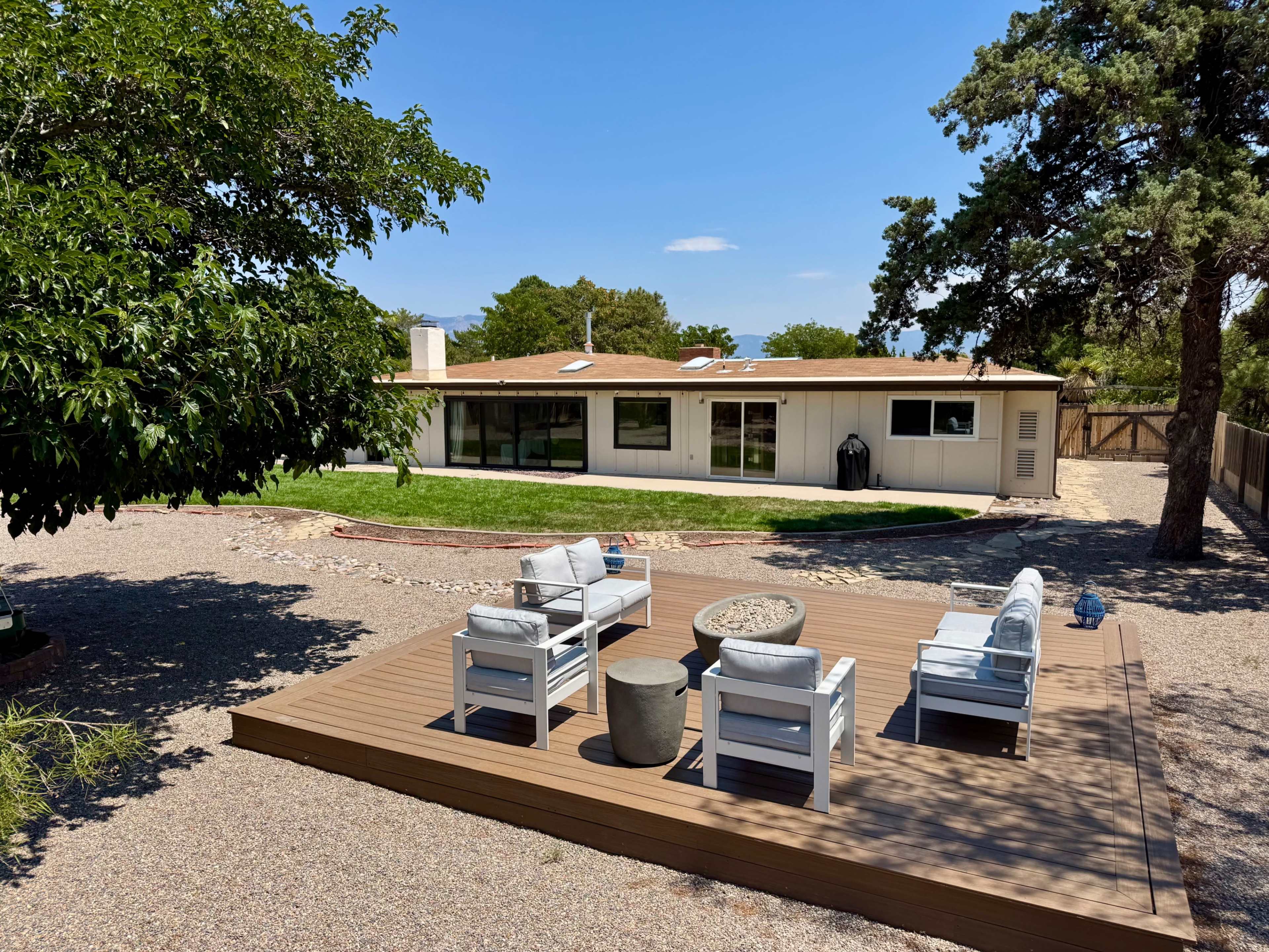 The image shows a backyard with a wooden deck featuring chairs and a table, overlooking a modern single-story house and a grassy area.