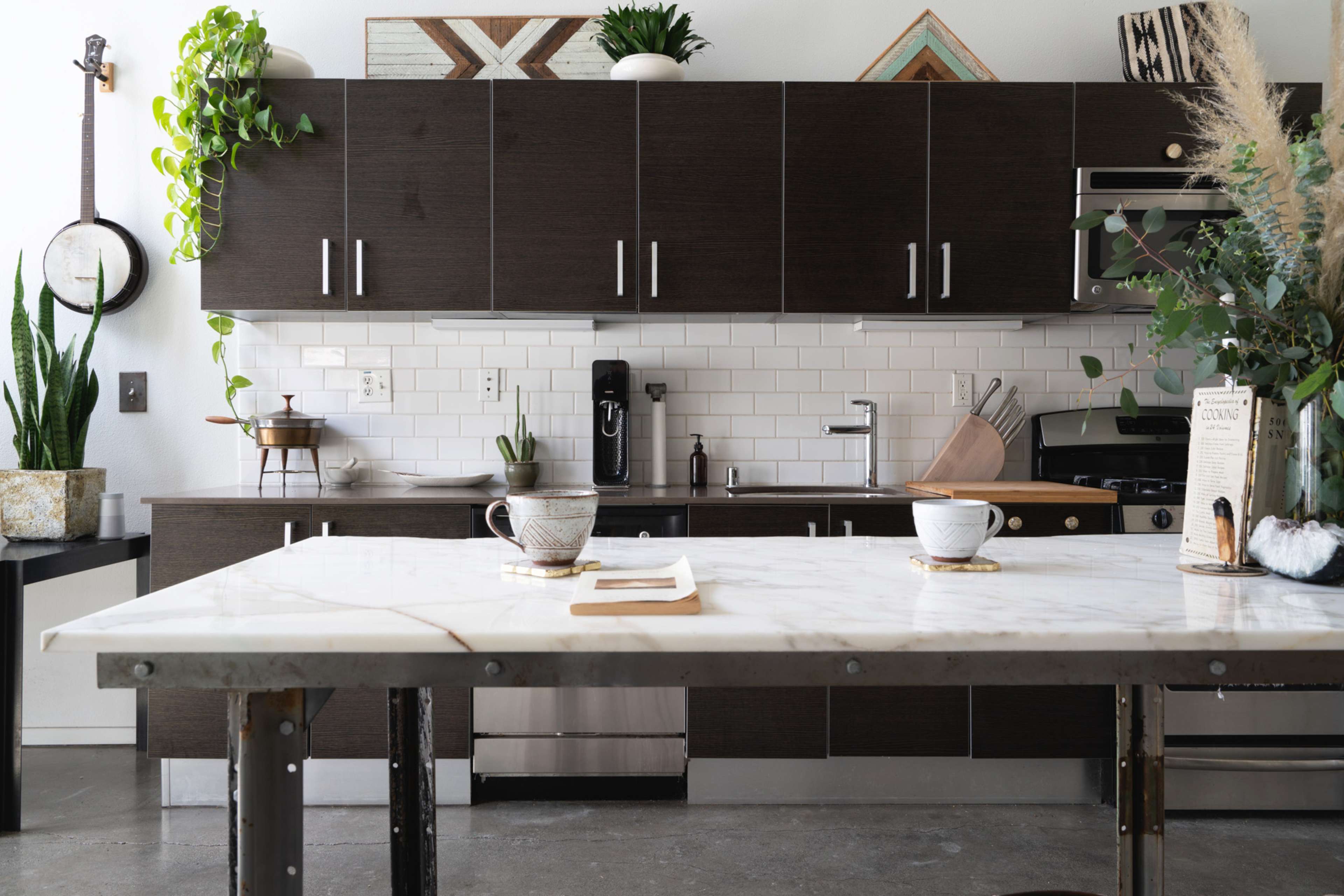 A modern kitchen features dark cabinetry, white subway tile, and a large marble-topped island with two coffee cups and an open book.