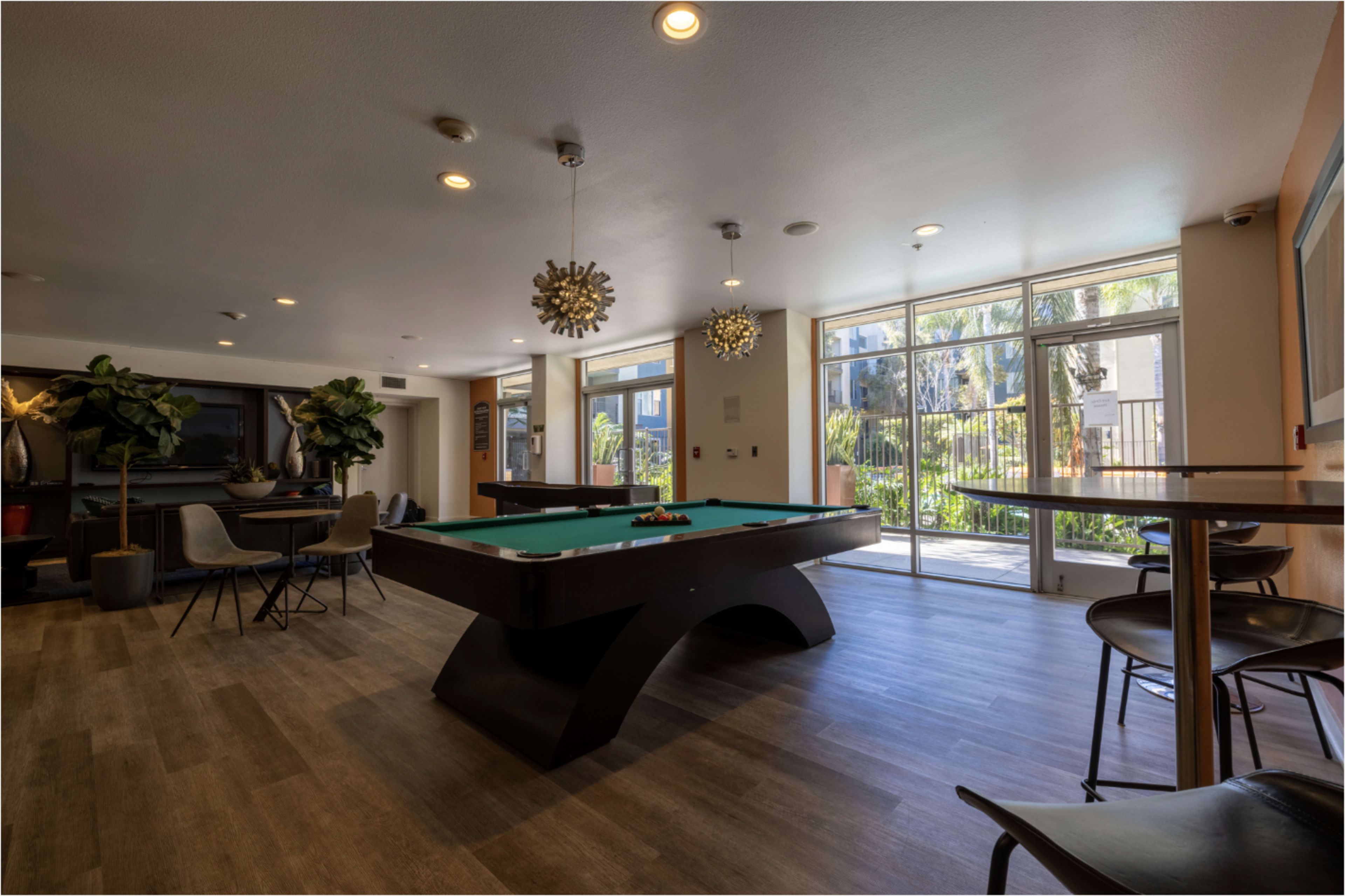 The image shows a modern lounge area featuring a billiards table, seating, and large windows overlooking an outdoor space with greenery.