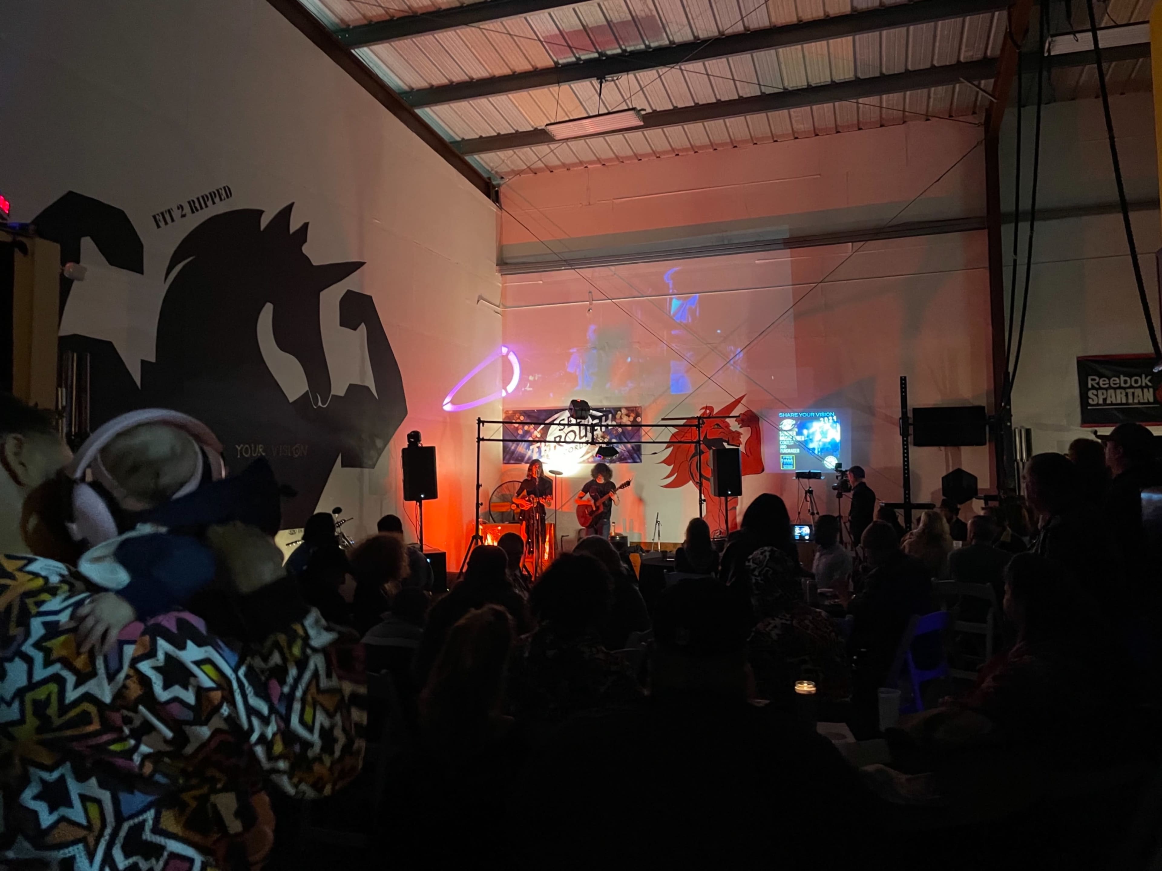 A crowd gathers in a dimly lit space to watch a live music performance, illuminated by colorful stage lights and a projection on the wall.