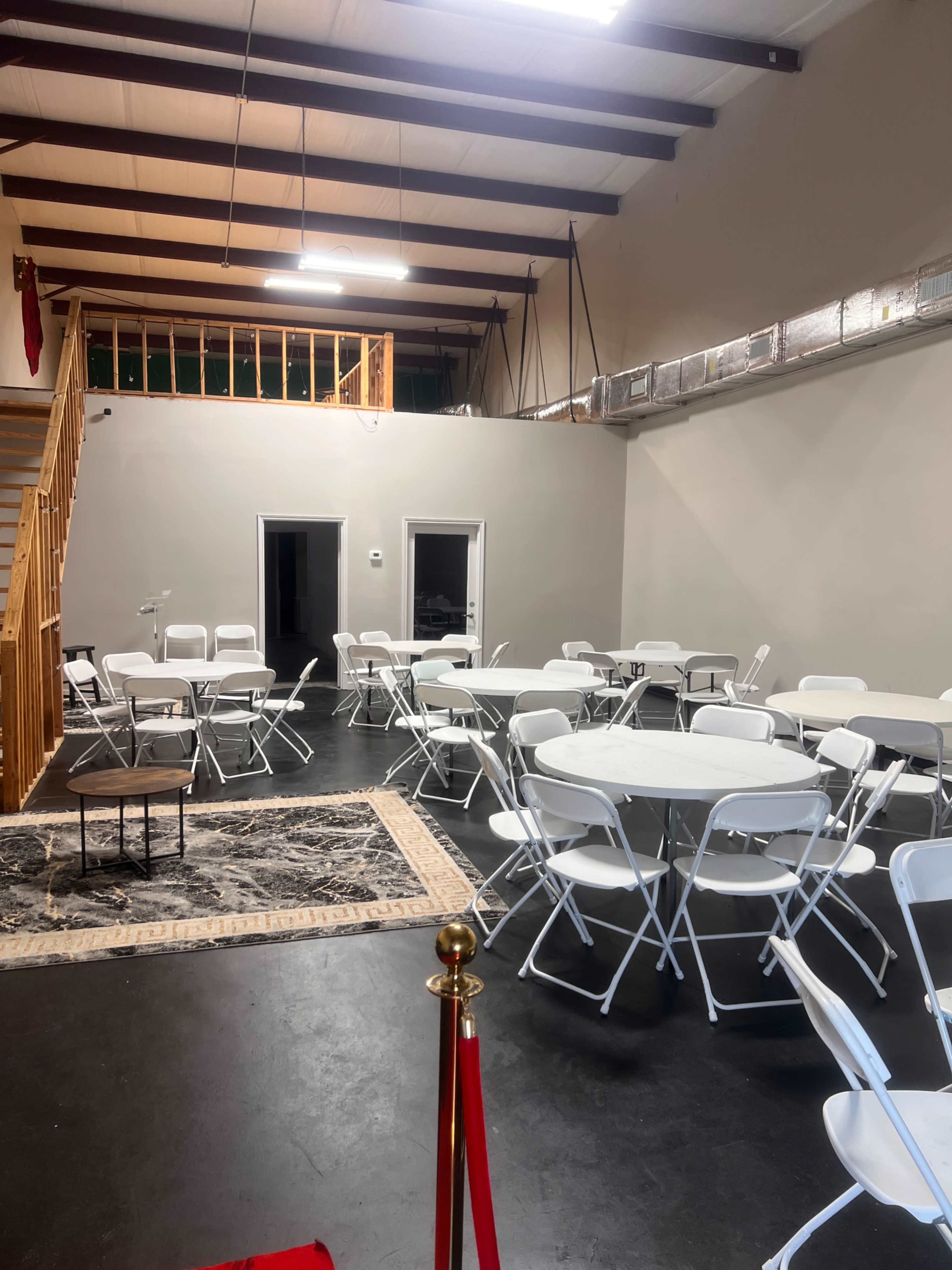 The image shows a spacious event area with white round tables and chairs arranged throughout, featuring a staircase in the background and two doorways on one side.