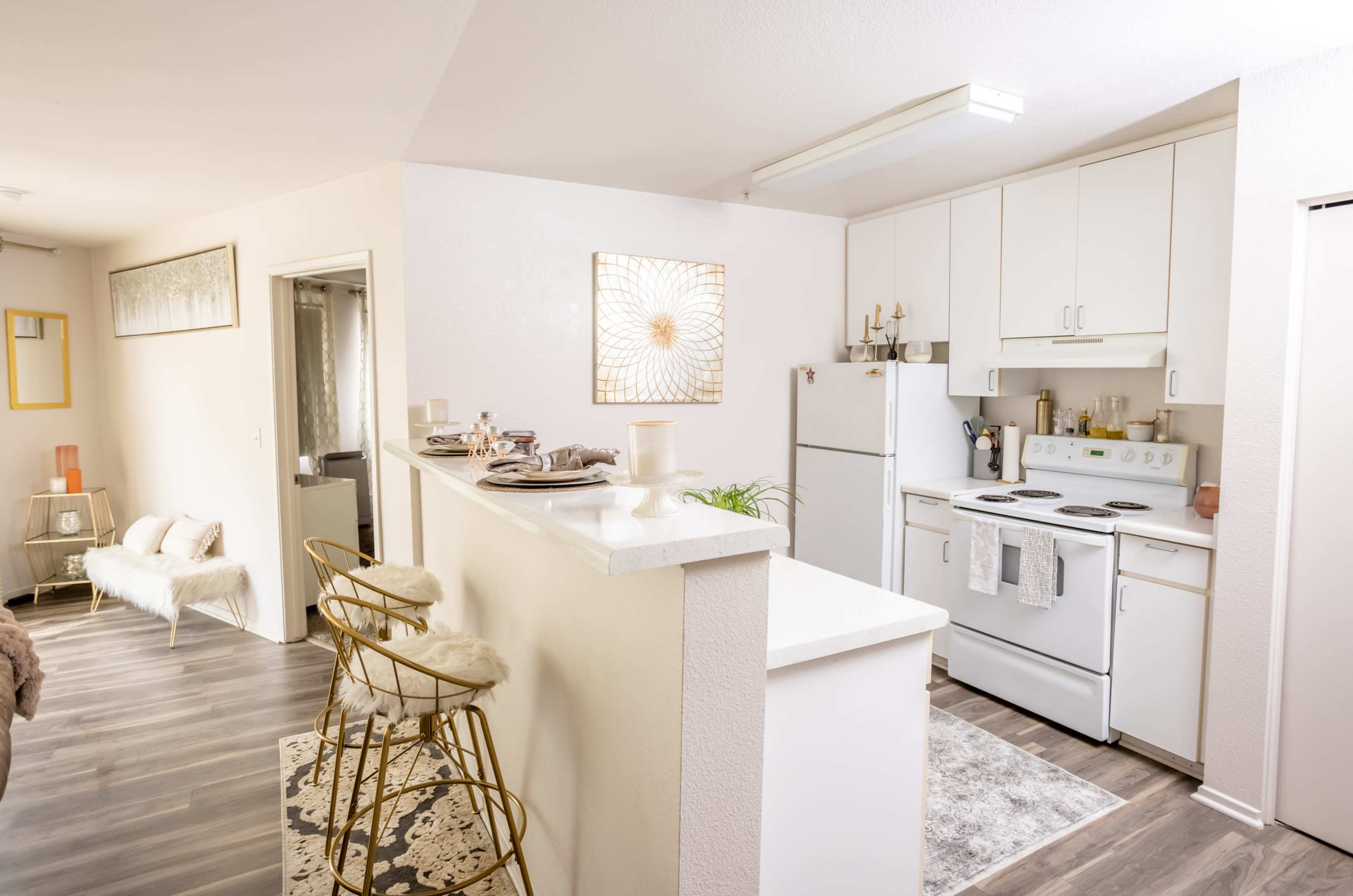 A modern kitchen features white cabinetry, an oven, a refrigerator, and bar seating with gold stools.