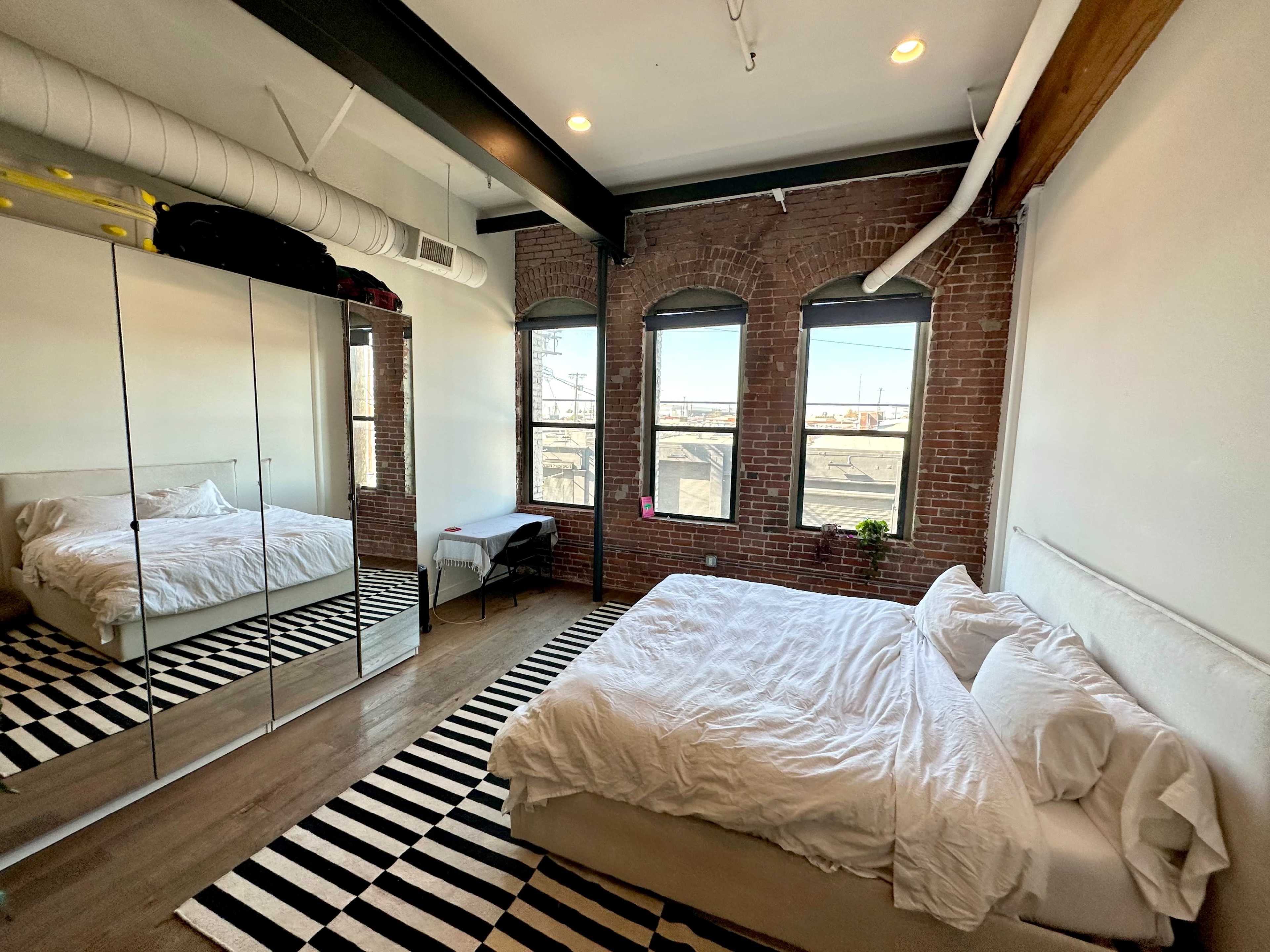 The bedroom features a large bed, a mirrored closet reflecting the room, and three arched windows with city views.