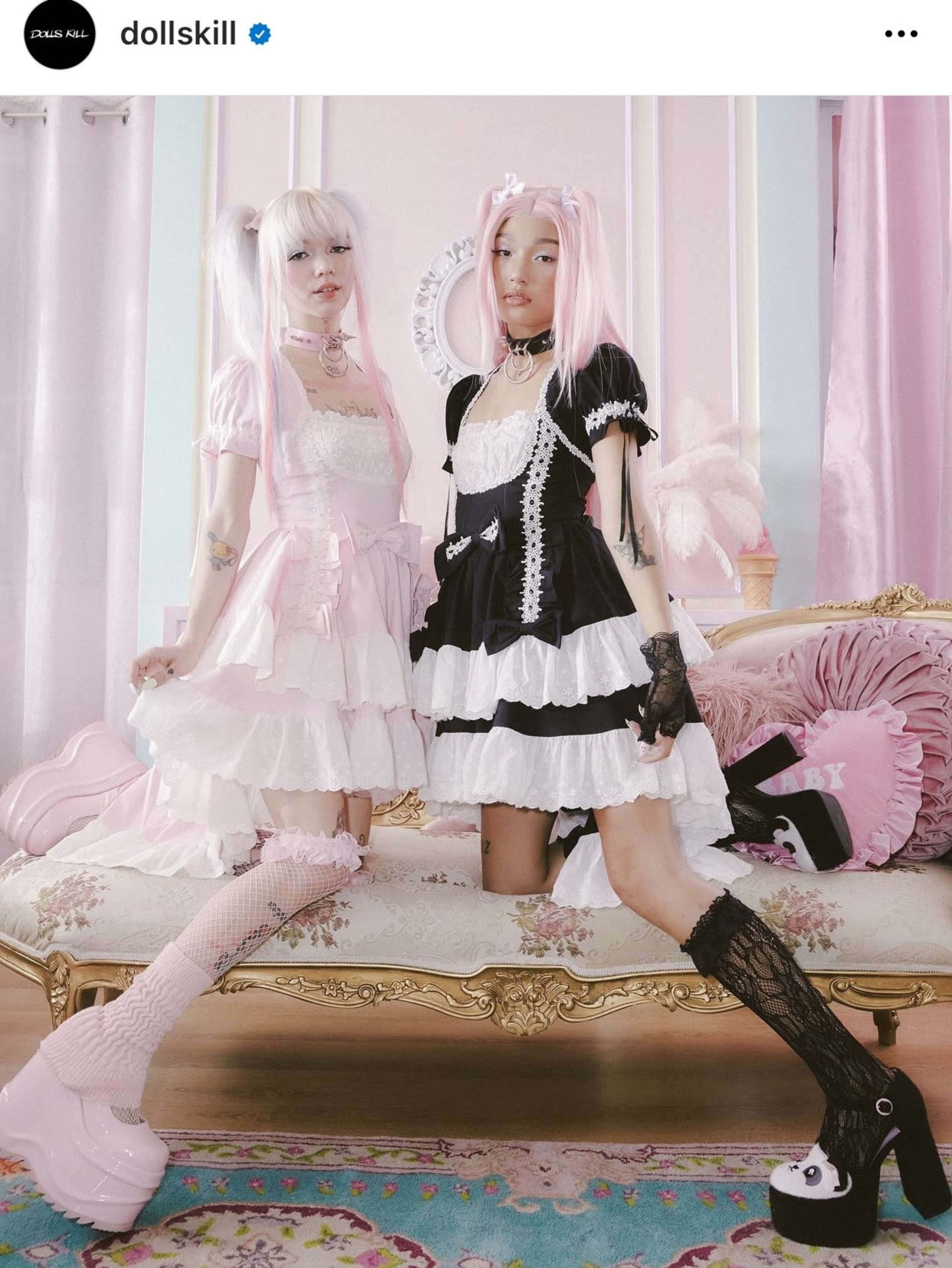 Two individuals in contrasting pastel-colored dresses stand in a decorated room with a vintage couch and soft lighting.