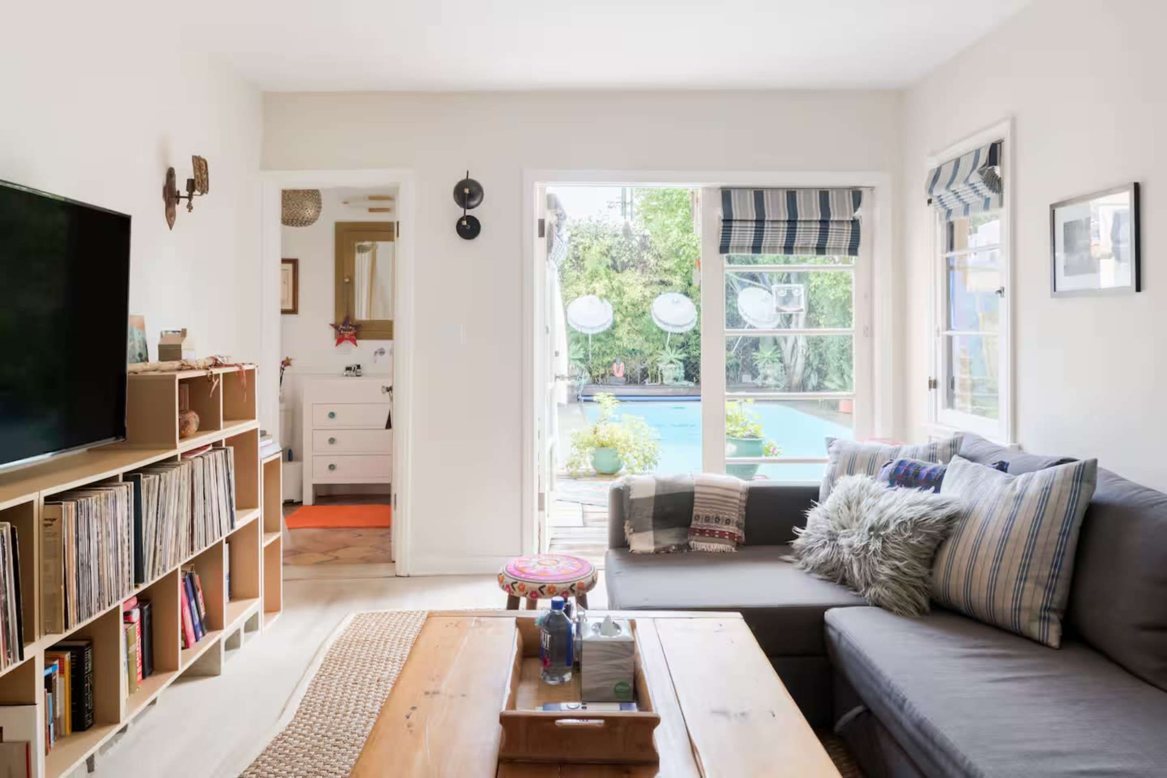 The living room features a gray sectional sofa, a wooden coffee table, and a large TV, with windows that provide views of a pool and greenery outside.