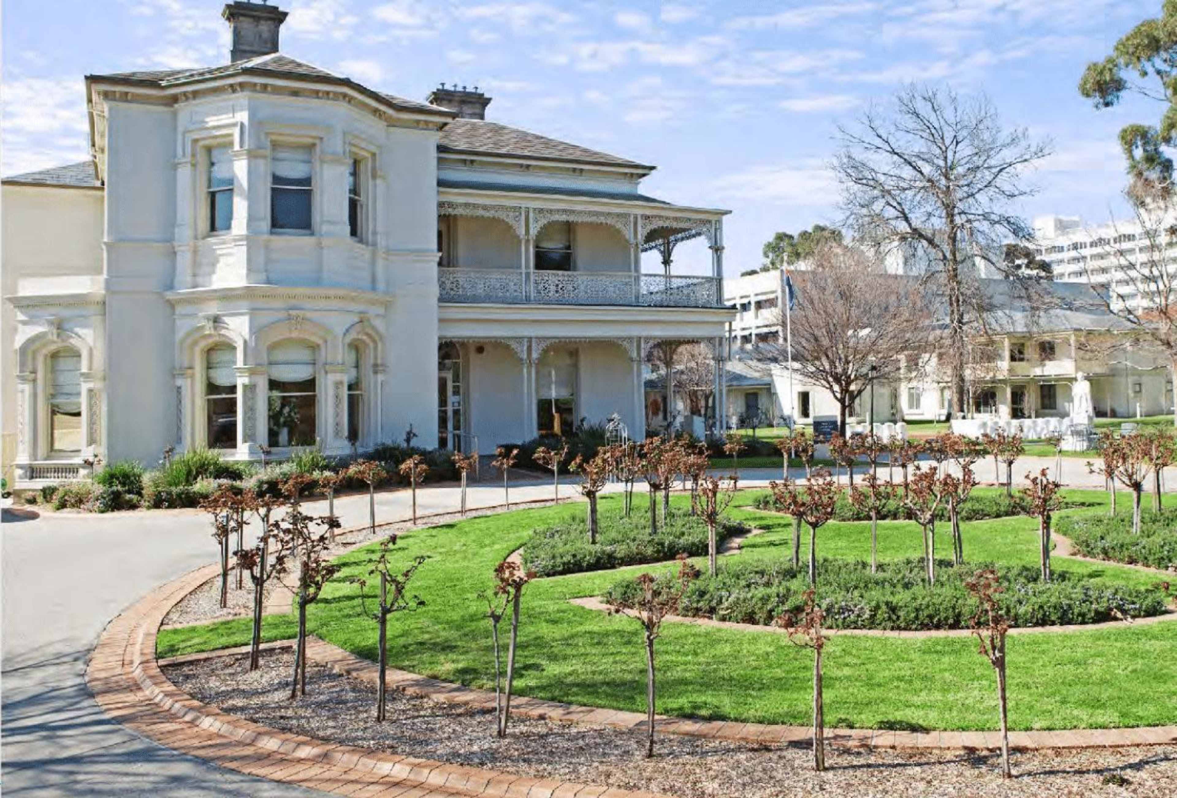 A large Victorian-style house stands beside a circular garden featuring symmetrical rows of small, leafless bushes.