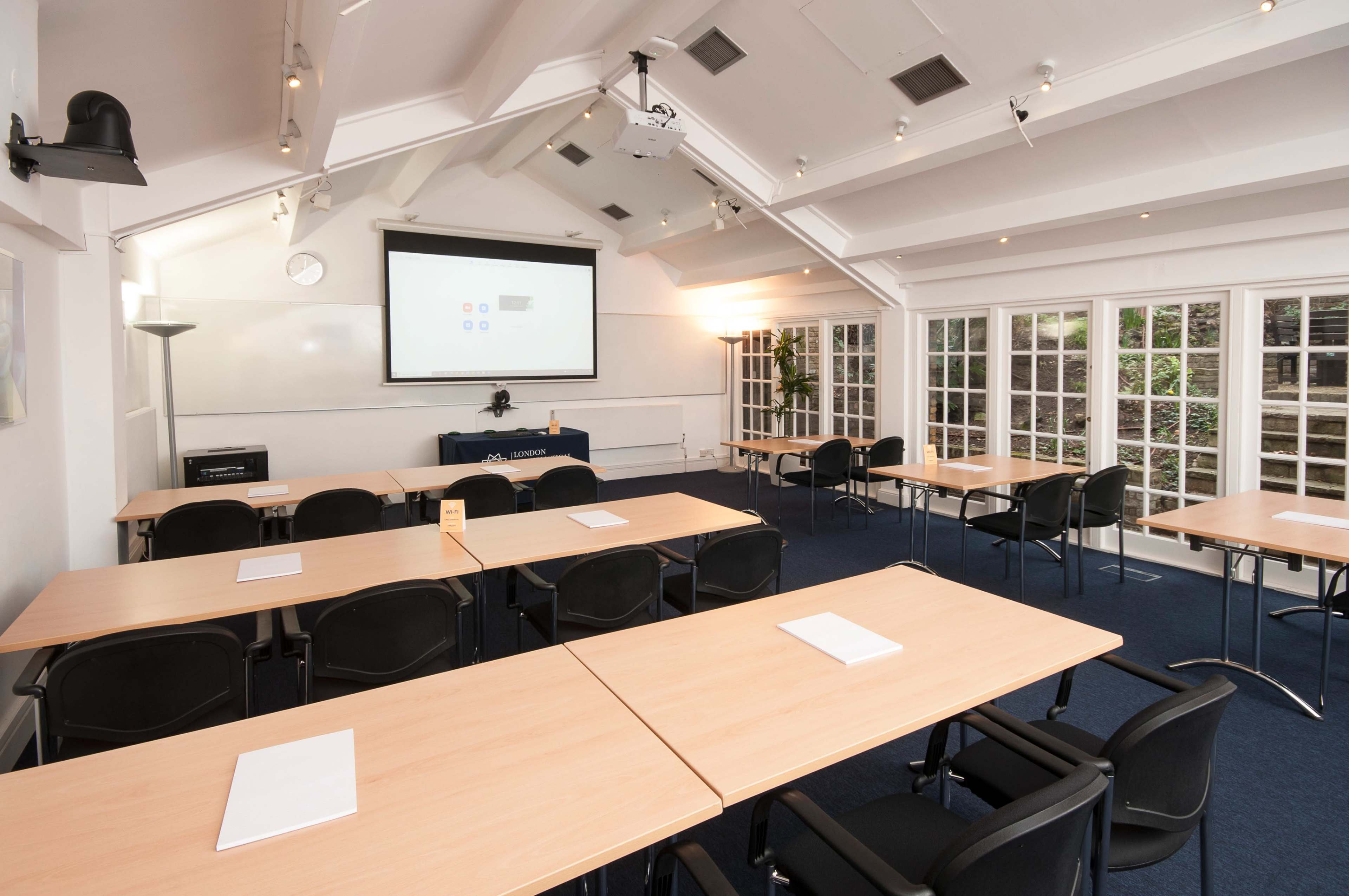 A conference room with several rectangular tables set up for a meeting, equipped with a projector screen at the front and large windows allowing natural light.
