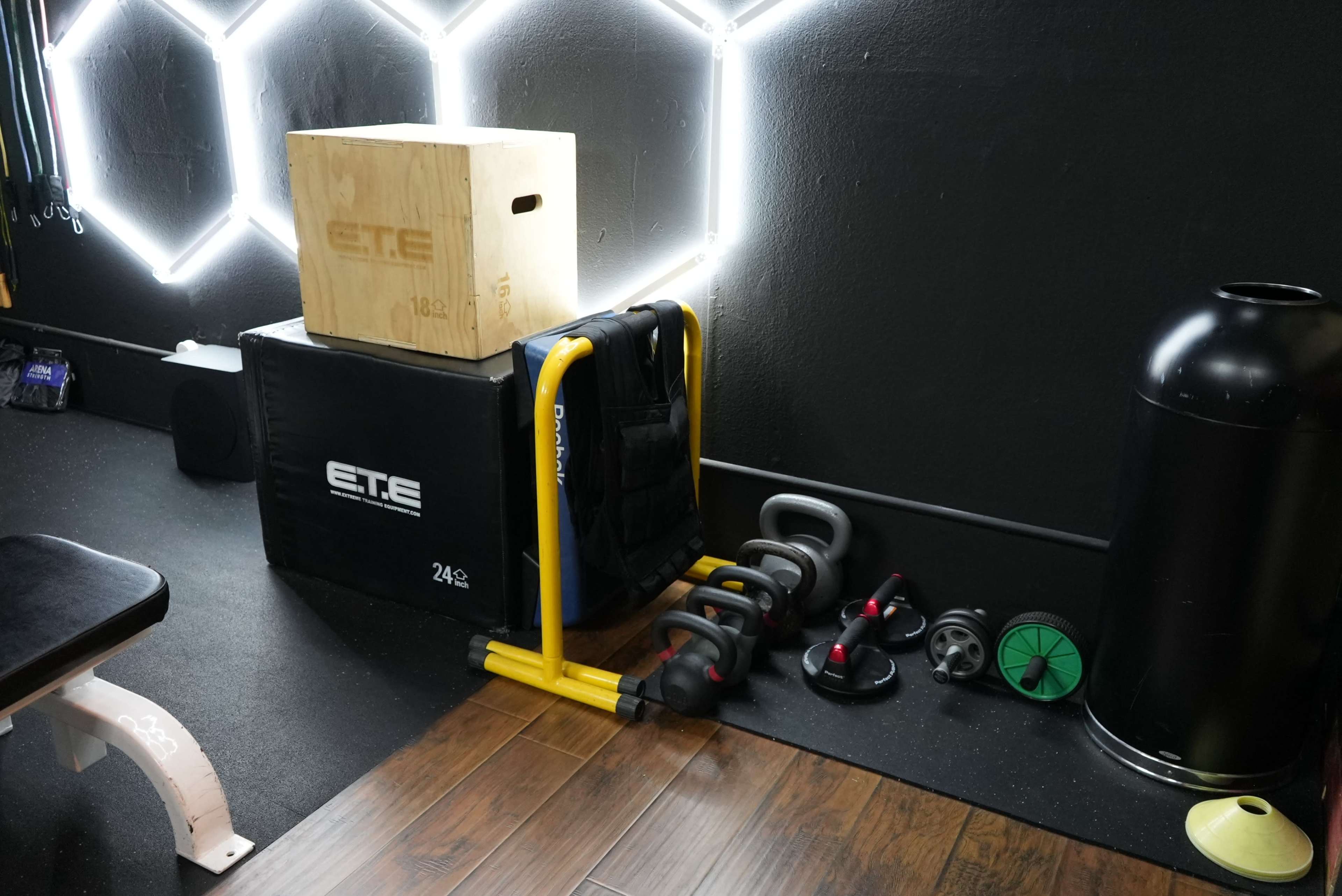 A fitness area with a wooden box, a padded black box, various dumbbells, and a black kettlebell on the floor, all illuminated by neon lights in the background.