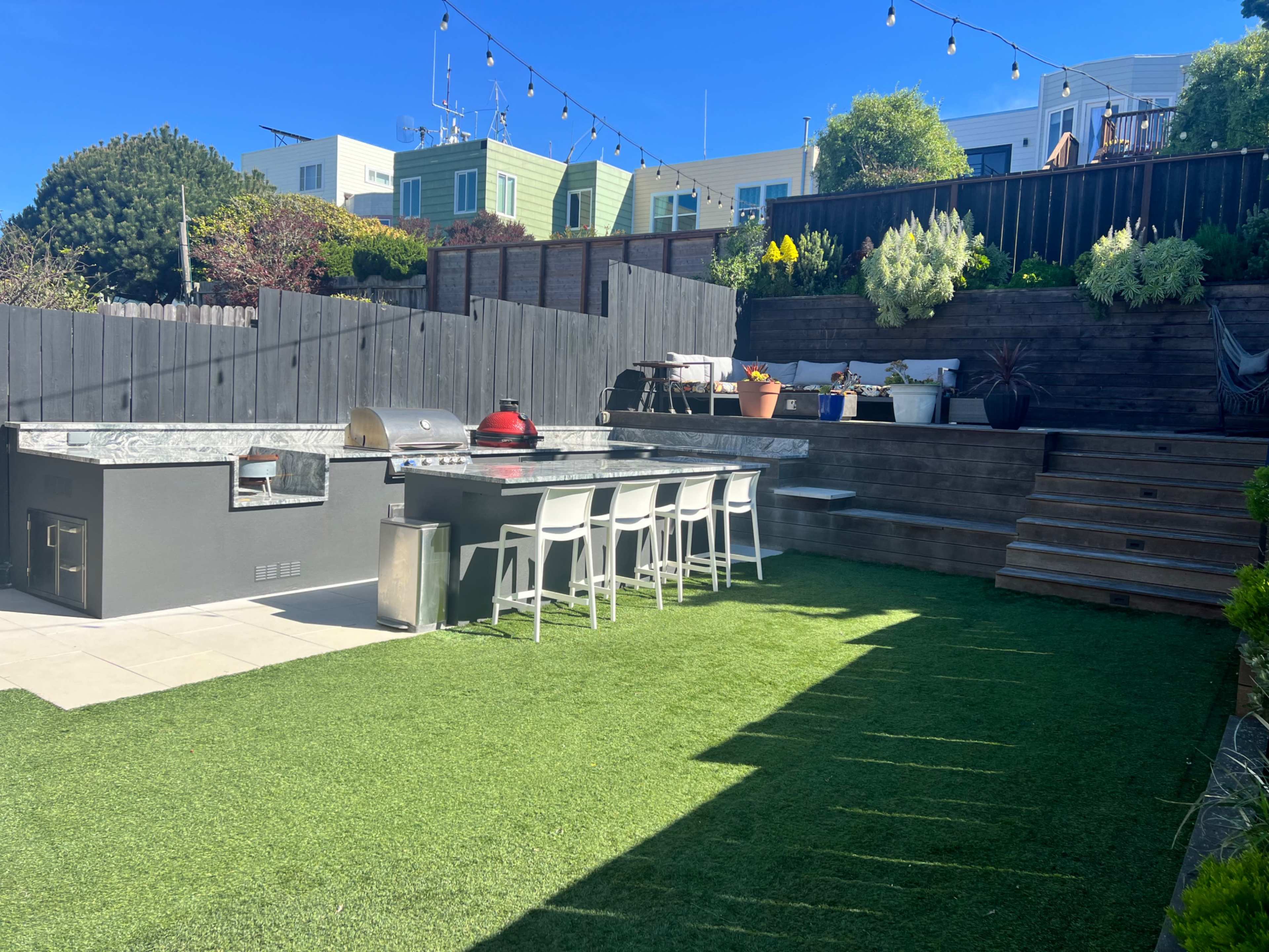 The image shows a modern outdoor kitchen area with a grill, seating for six, and a landscaped yard featuring artificial grass and steps leading to an elevated garden.