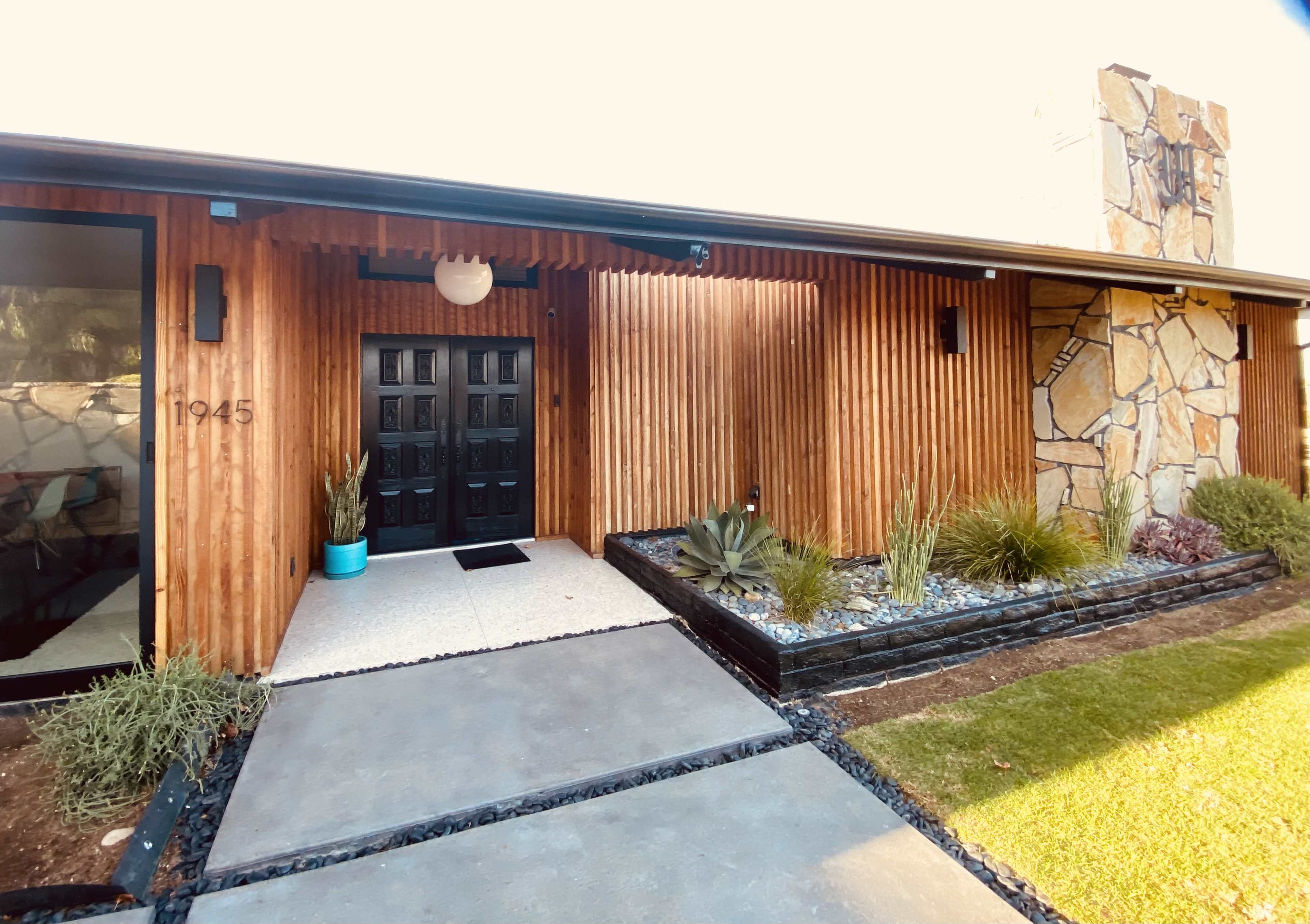 The image shows a modern home entrance with wooden siding, a stone feature wall, and a paved pathway leading to double black doors.