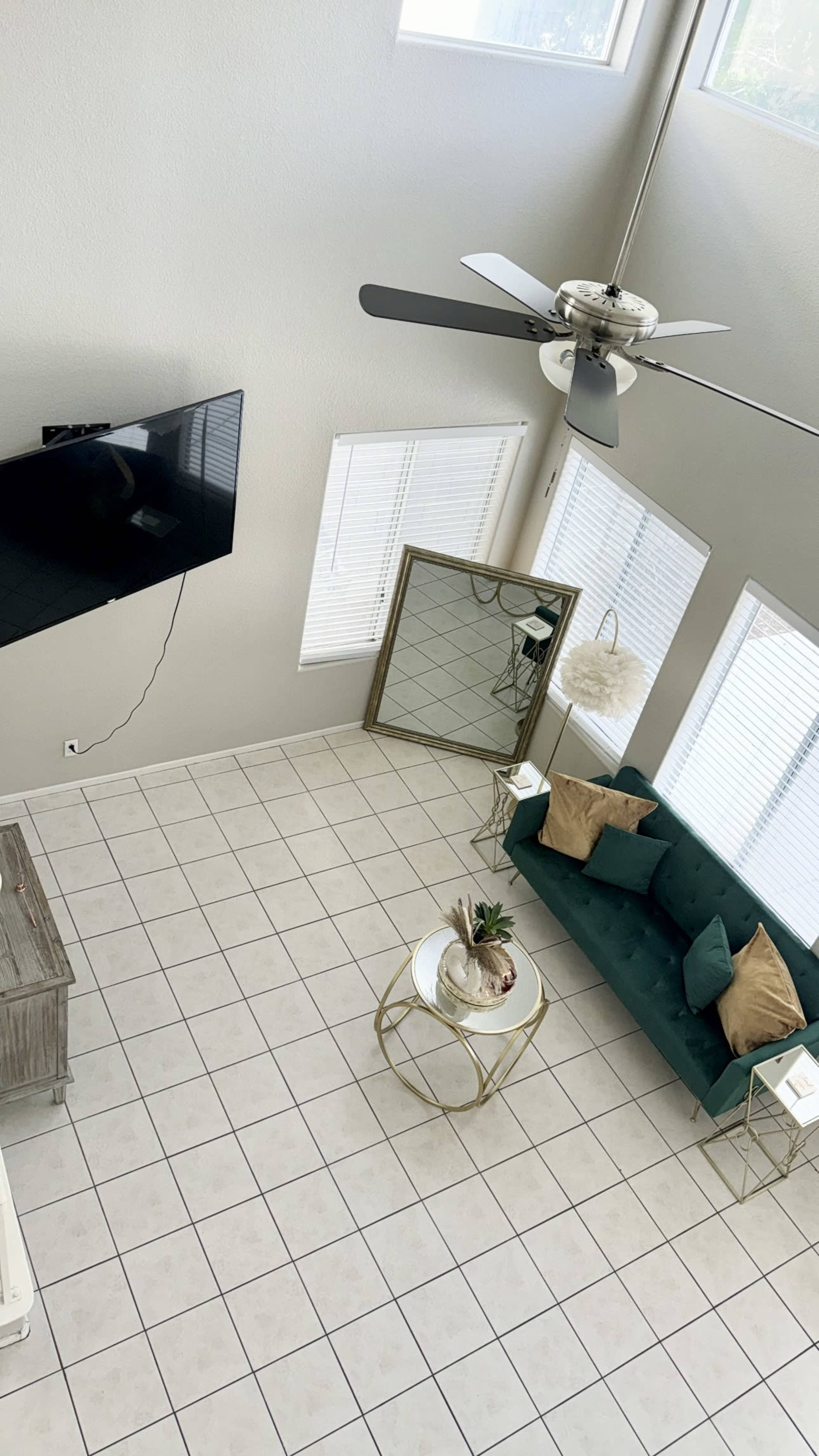 The living room features a green sofa, a round coffee table, and a large mirror against the wall, with a ceiling fan and large windows allowing natural light to enter.