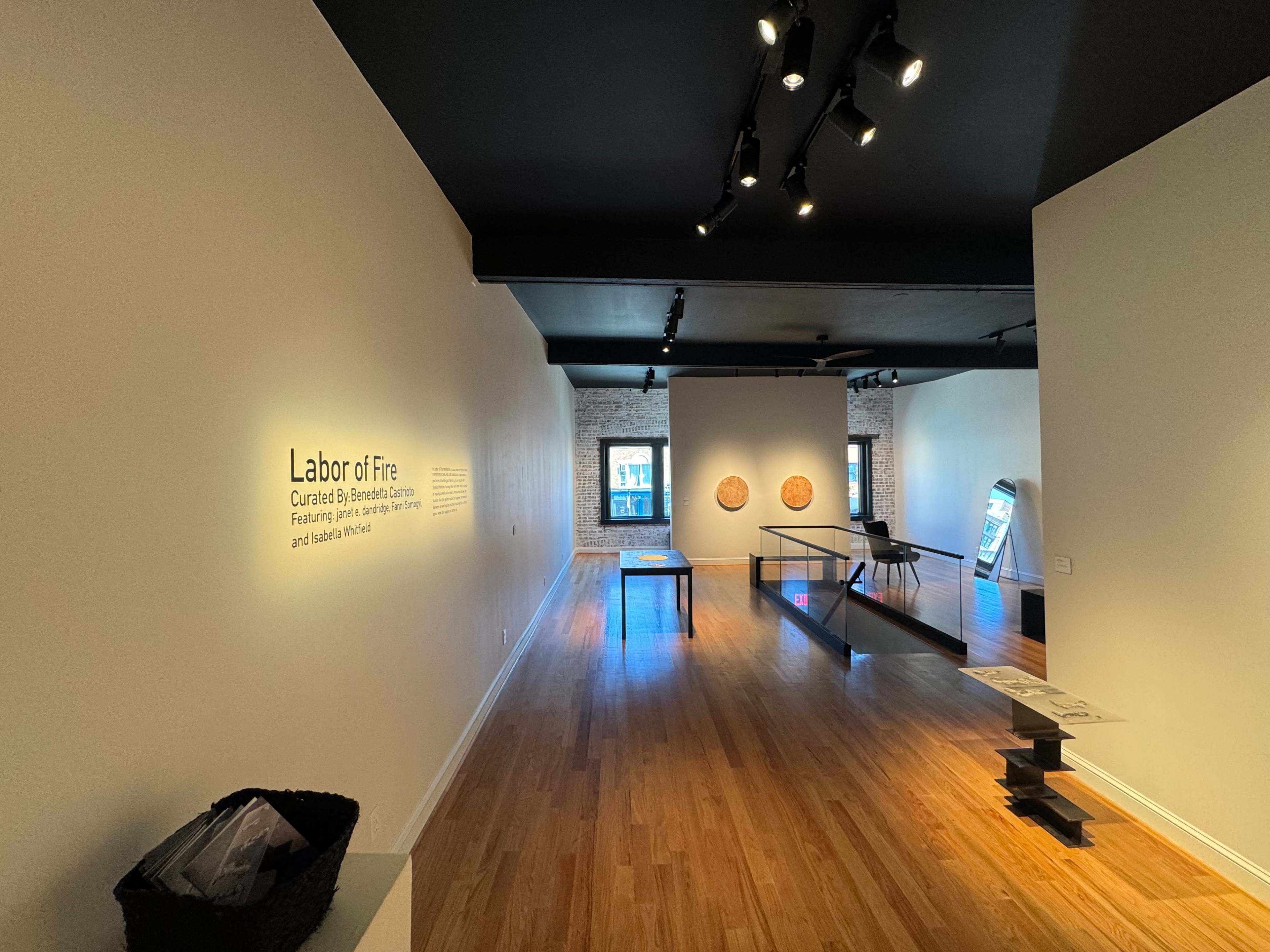 The image shows an art gallery with a white wall featuring an exhibition title, wooden artworks on the wall, and minimal furnishings in the space.
