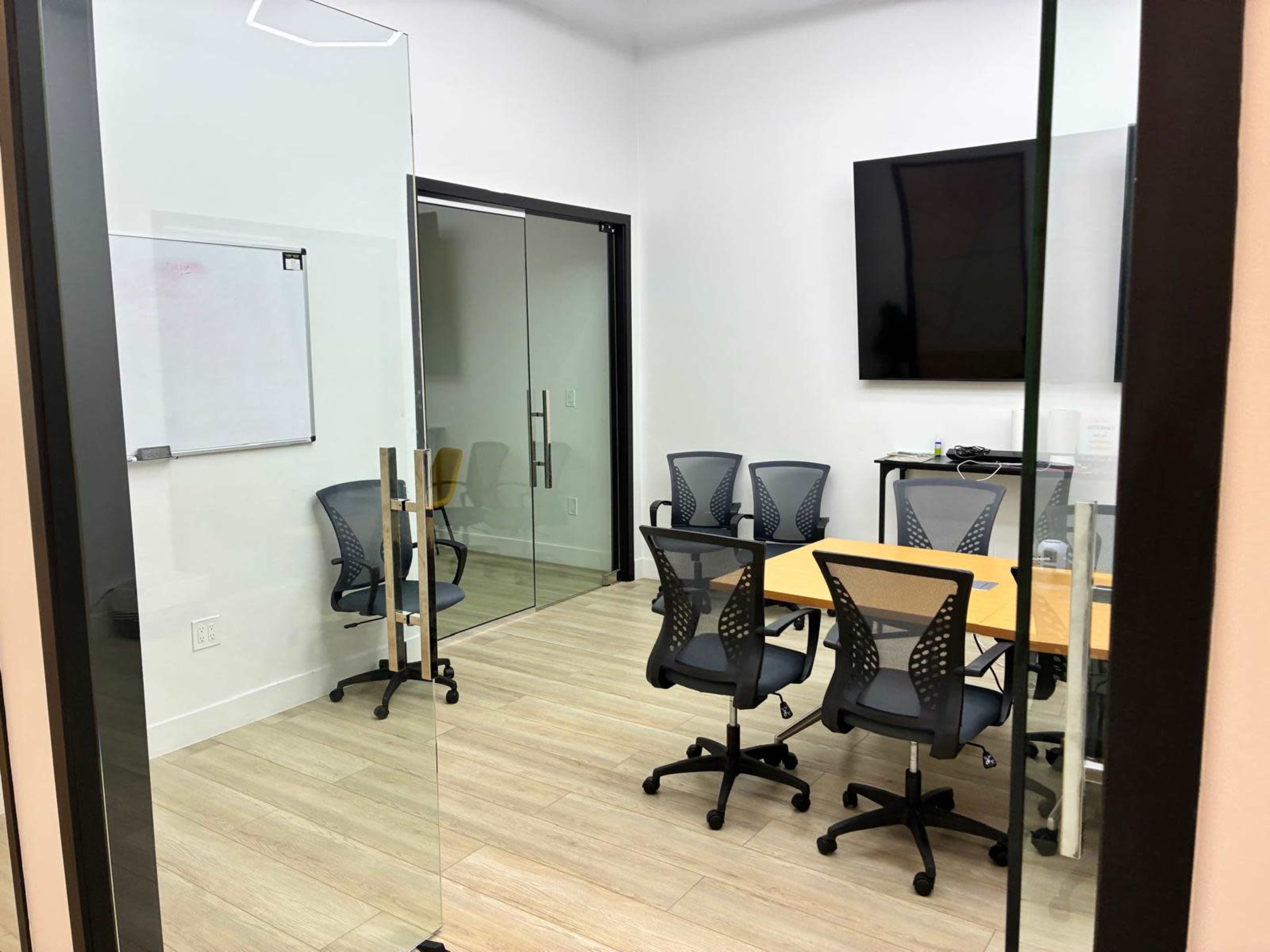 The image shows a modern conference room with a rectangular wooden table, six ergonomic chairs, a whiteboard, and a screen mounted on the wall.