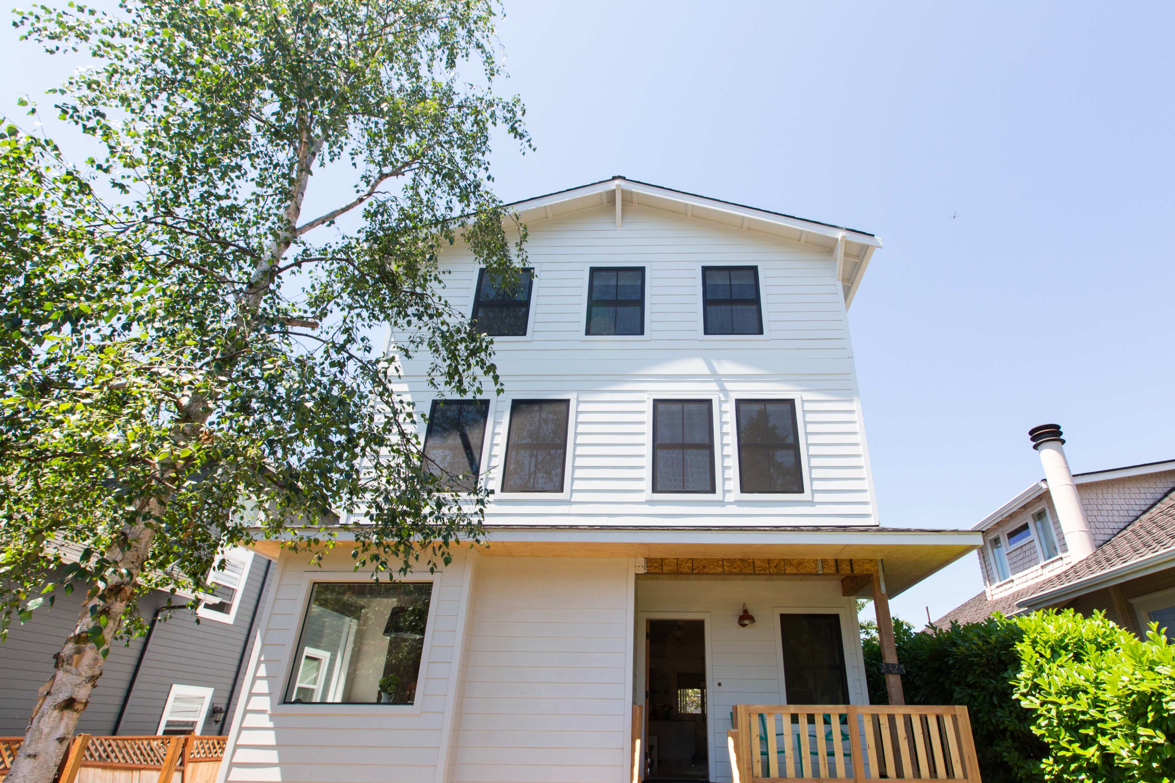 A three-story white house with multiple windows and a wooden front porch is surrounded by greenery and a birch tree.
