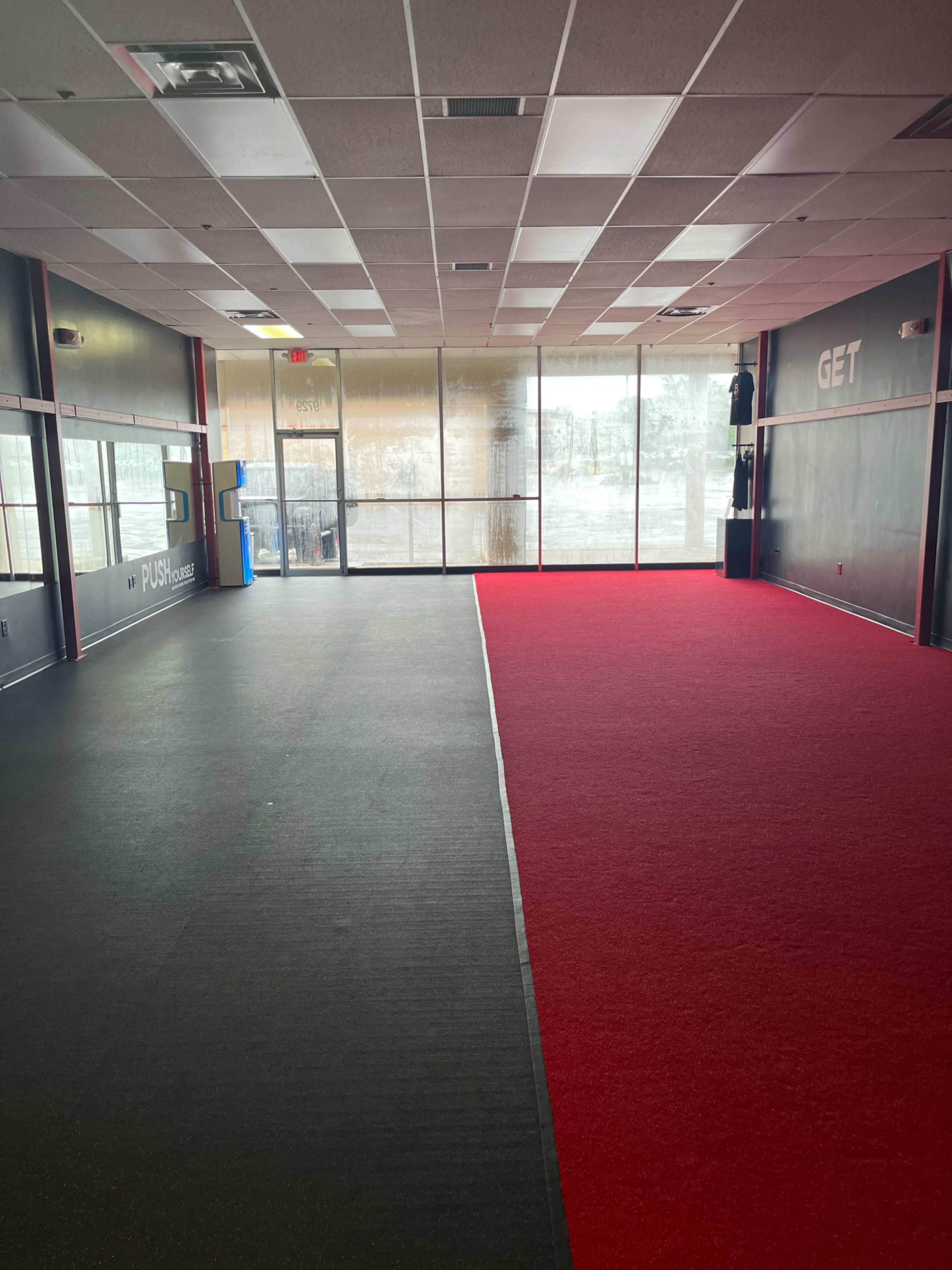 A spacious gym interior, featuring a red carpeted area on one side and a black rubber floor on the other, with large windows providing natural light.