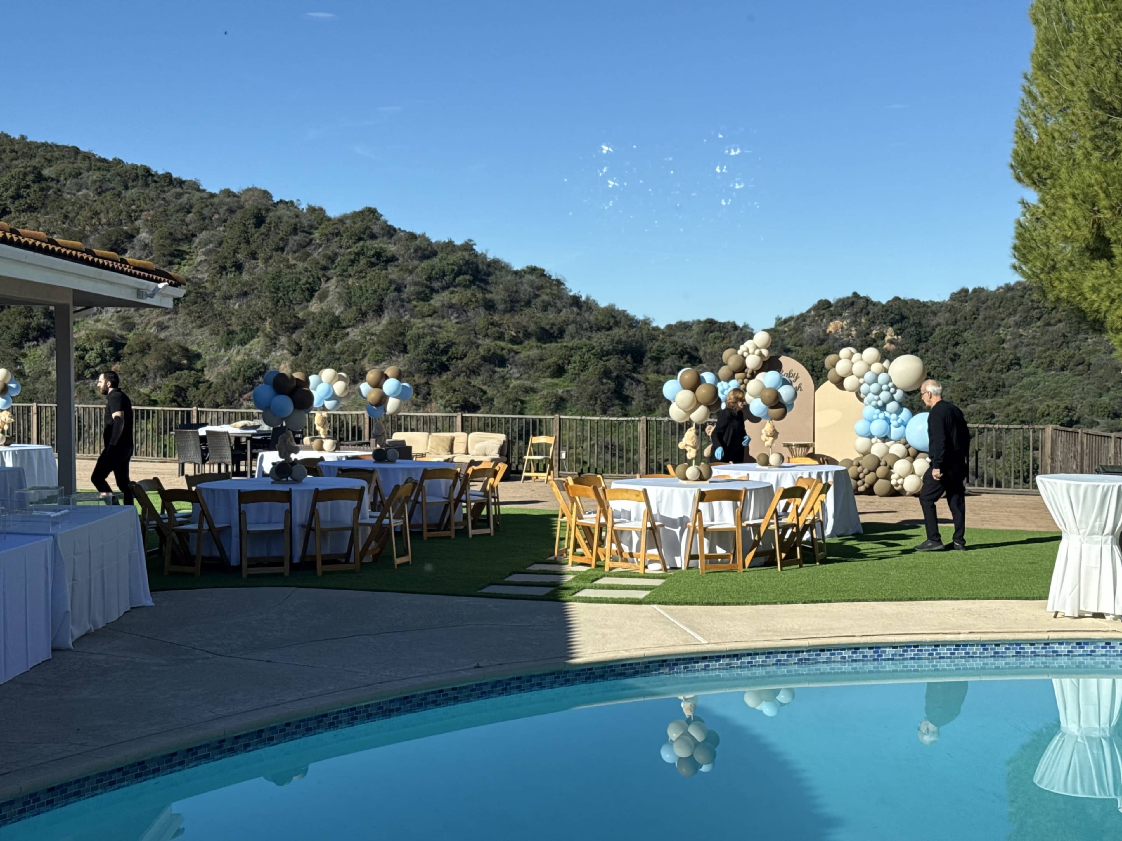 A landscaped outdoor area features tables decorated for an event, a pool reflecting the sky, and party decorations with balloons in blue and beige.