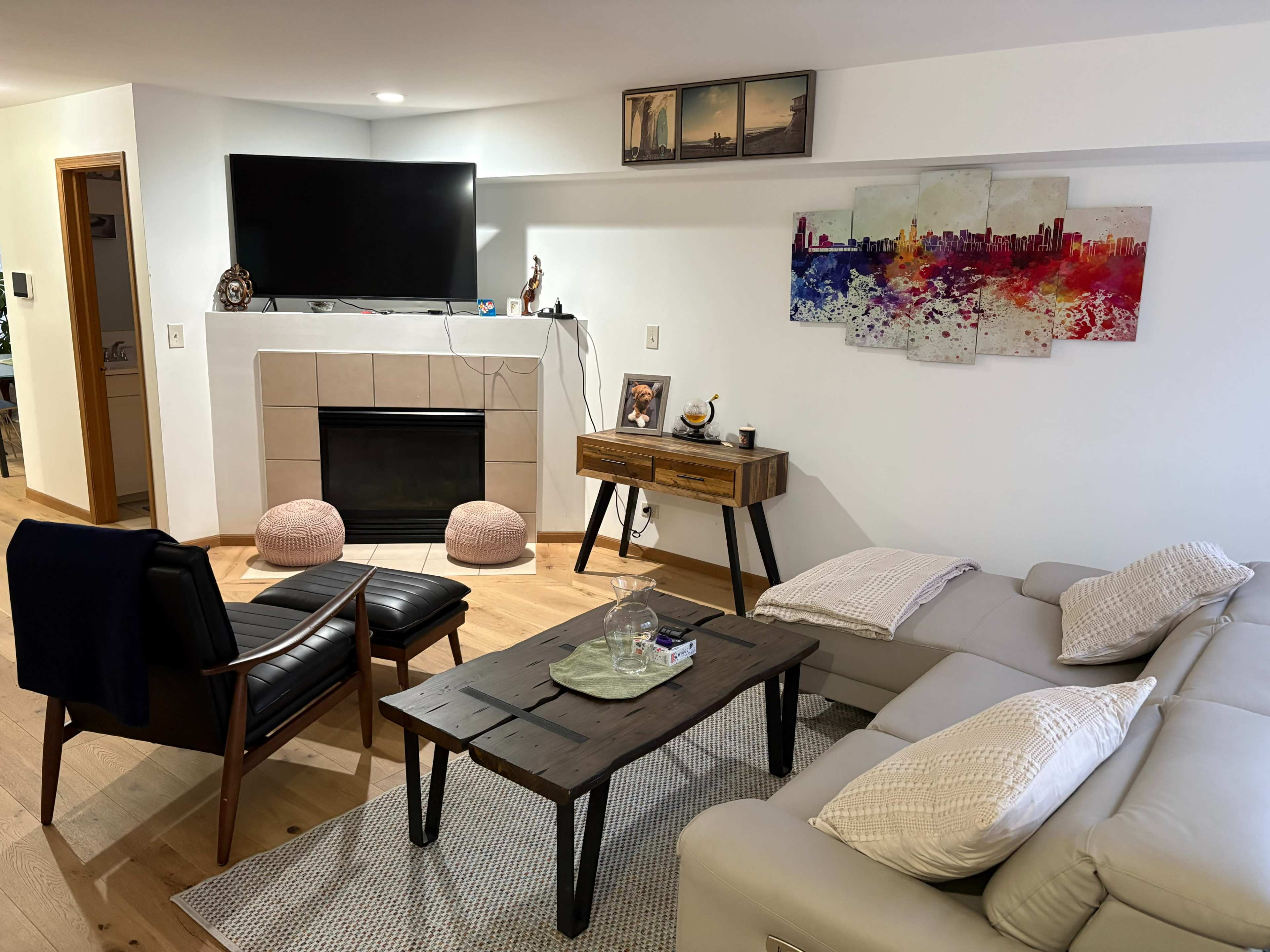 The living room features a large television mounted on the wall above a fireplace, with a minimalist couch, a wooden coffee table, and two poufs on a textured rug.