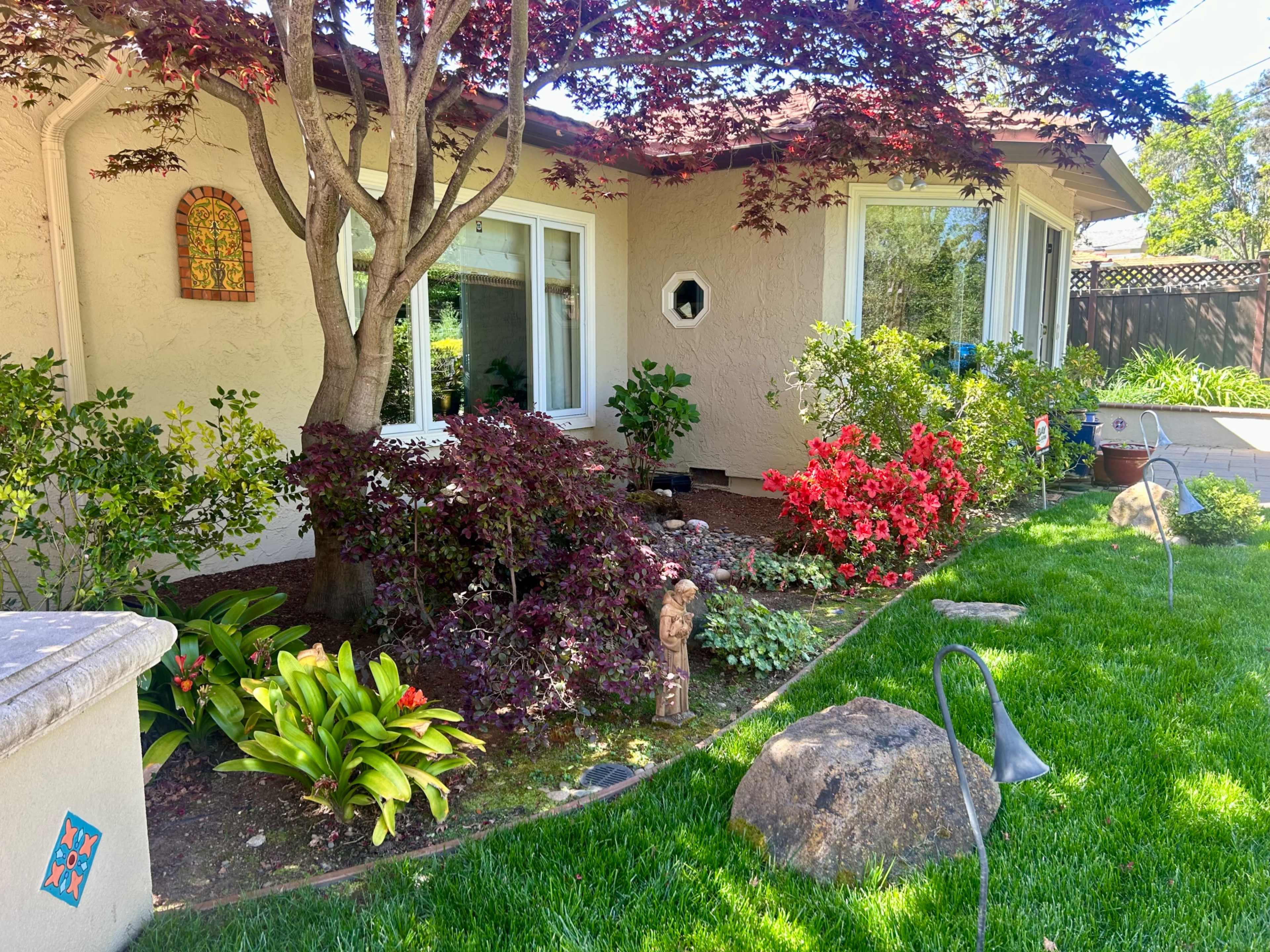 A garden in front of a house, featuring a variety of colorful plants, bushes, and a decorative stone statue near a tree.