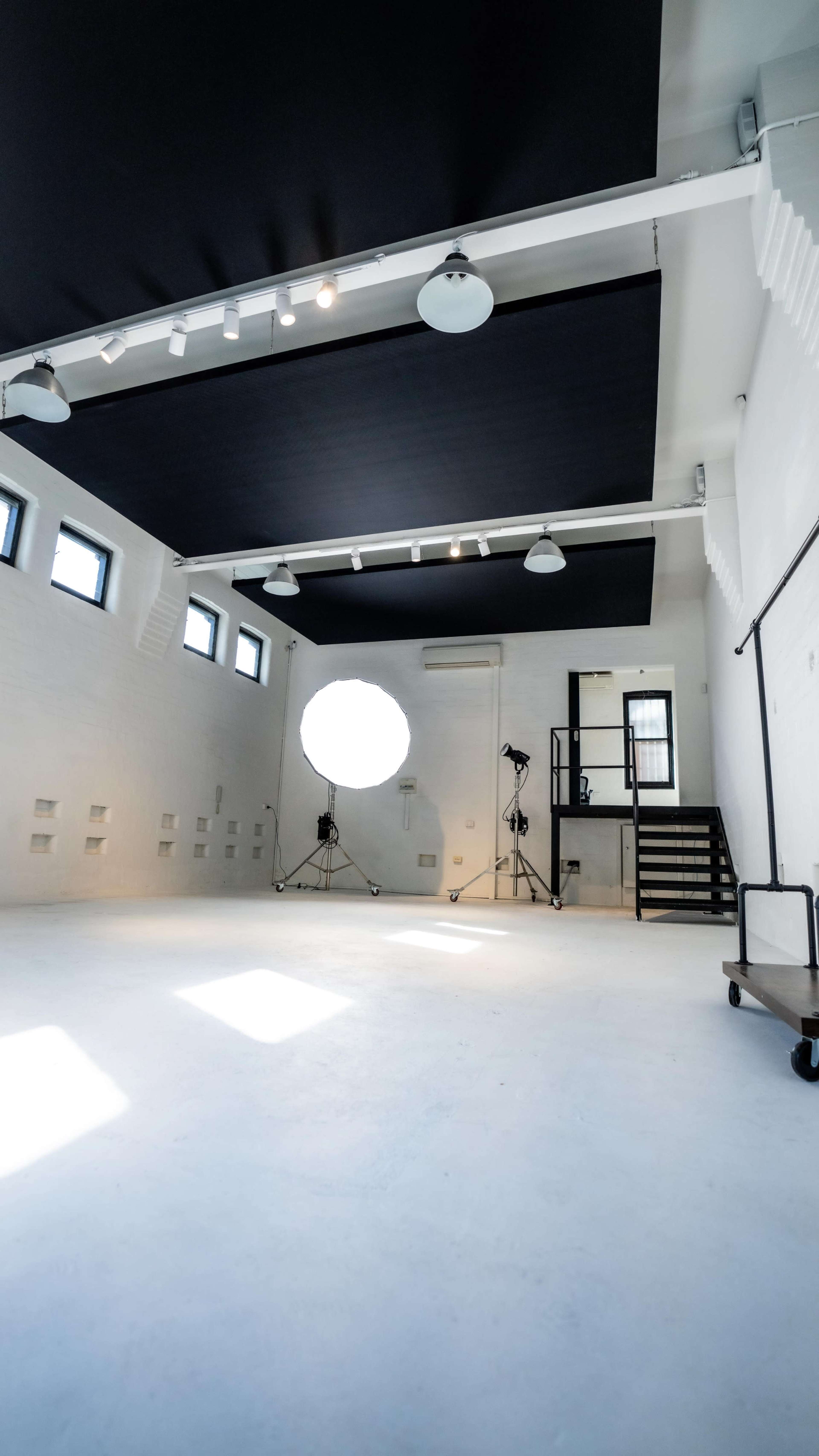 A spacious photography studio with a white floor, black overhead panels, and a large circular softbox on a stand.