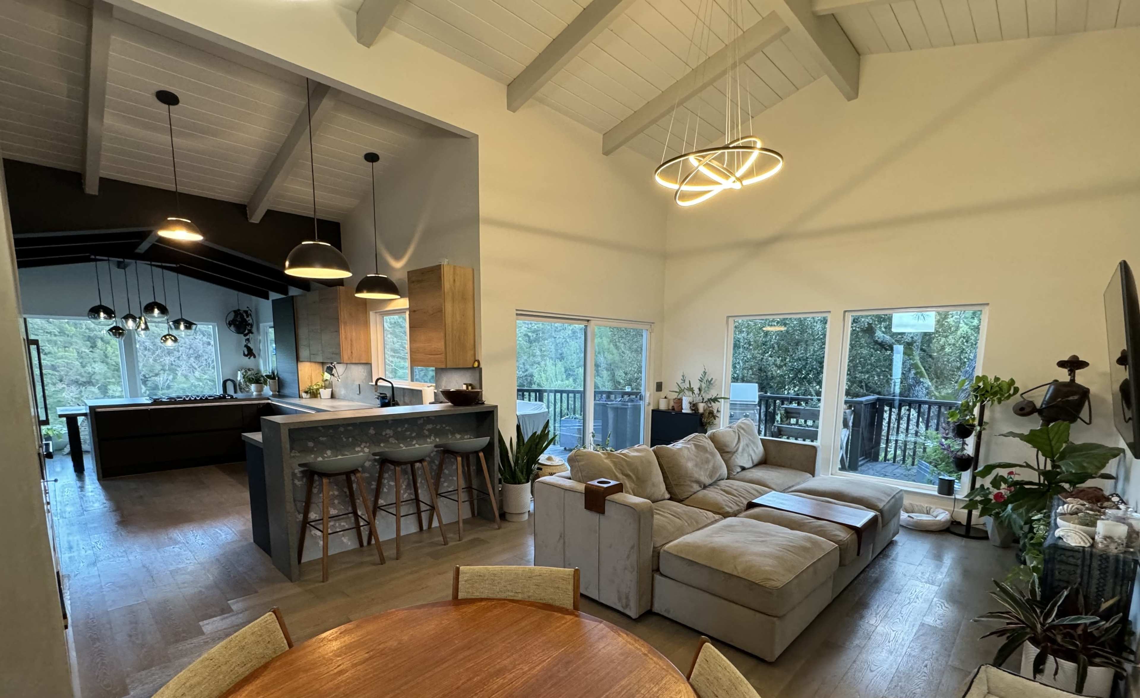 A modern open-concept living area with a beige sectional sofa, a wooden dining table, and a kitchen featuring dark cabinetry and pendant lighting.