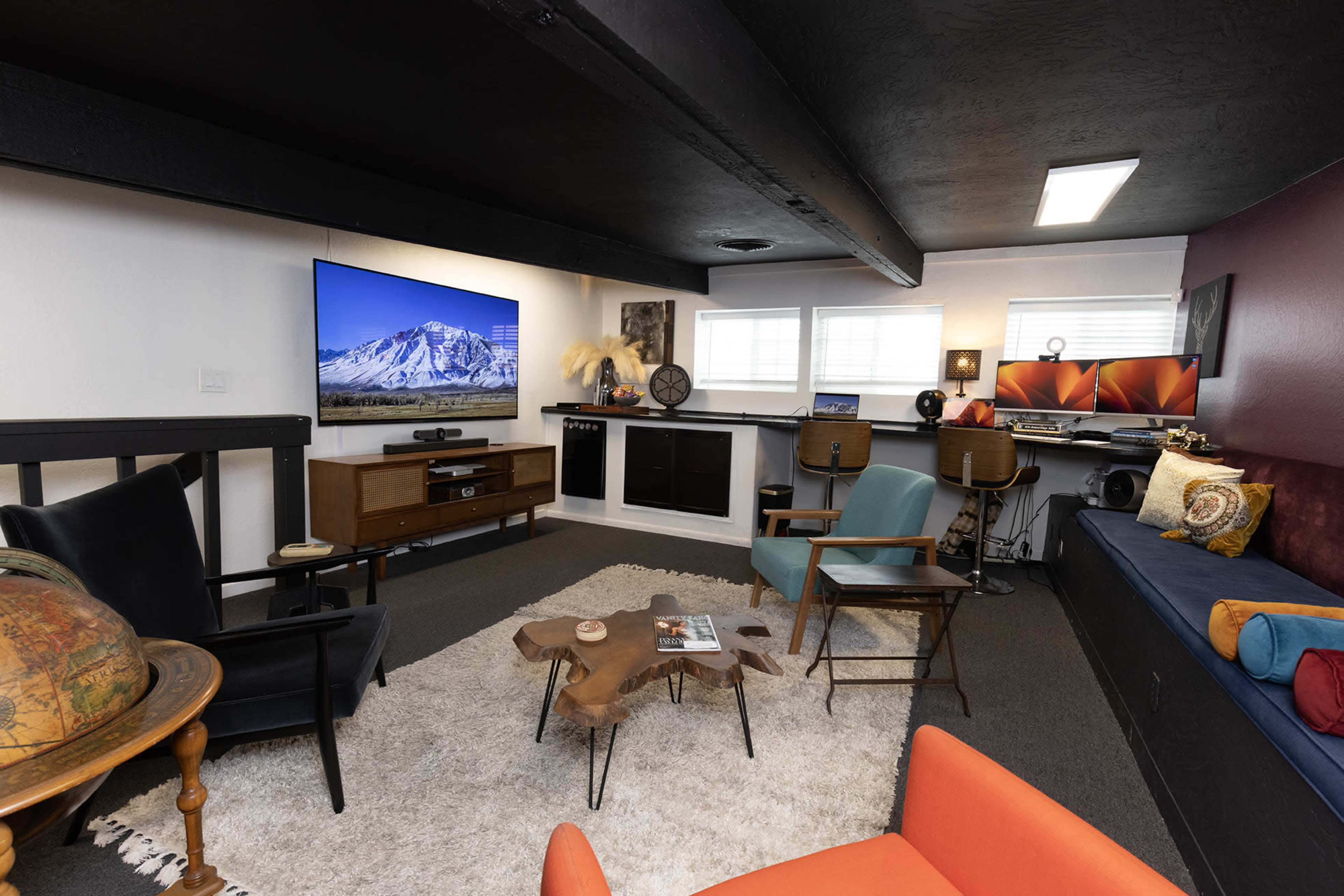 The image shows a cozy living space featuring a television, a computer setup, colorful armchairs, and a coffee table surrounded by a rug.