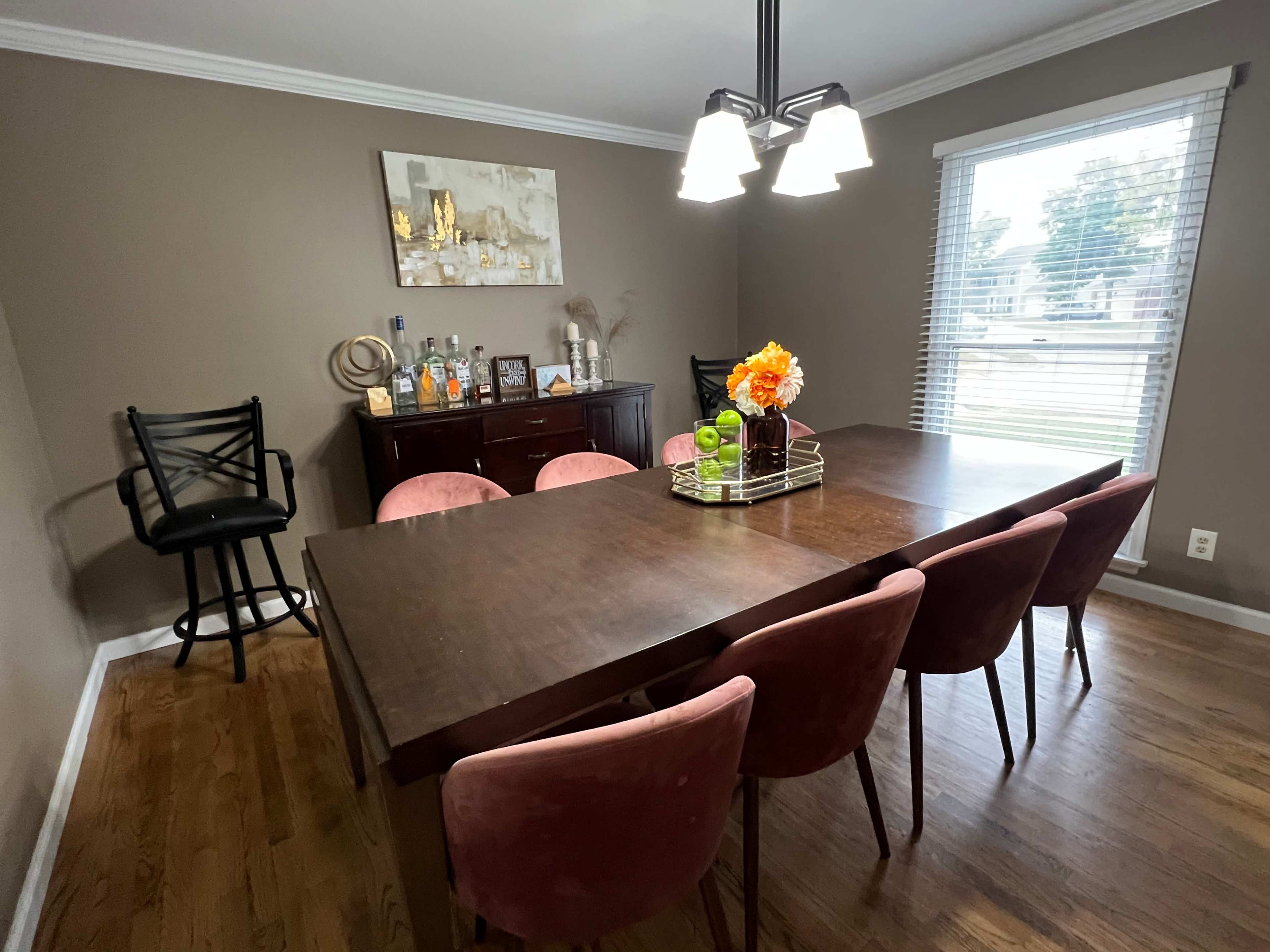 A dining room features a dark wooden table surrounded by pink chairs, with a bar cart and decorative items on a sideboard against the wall.