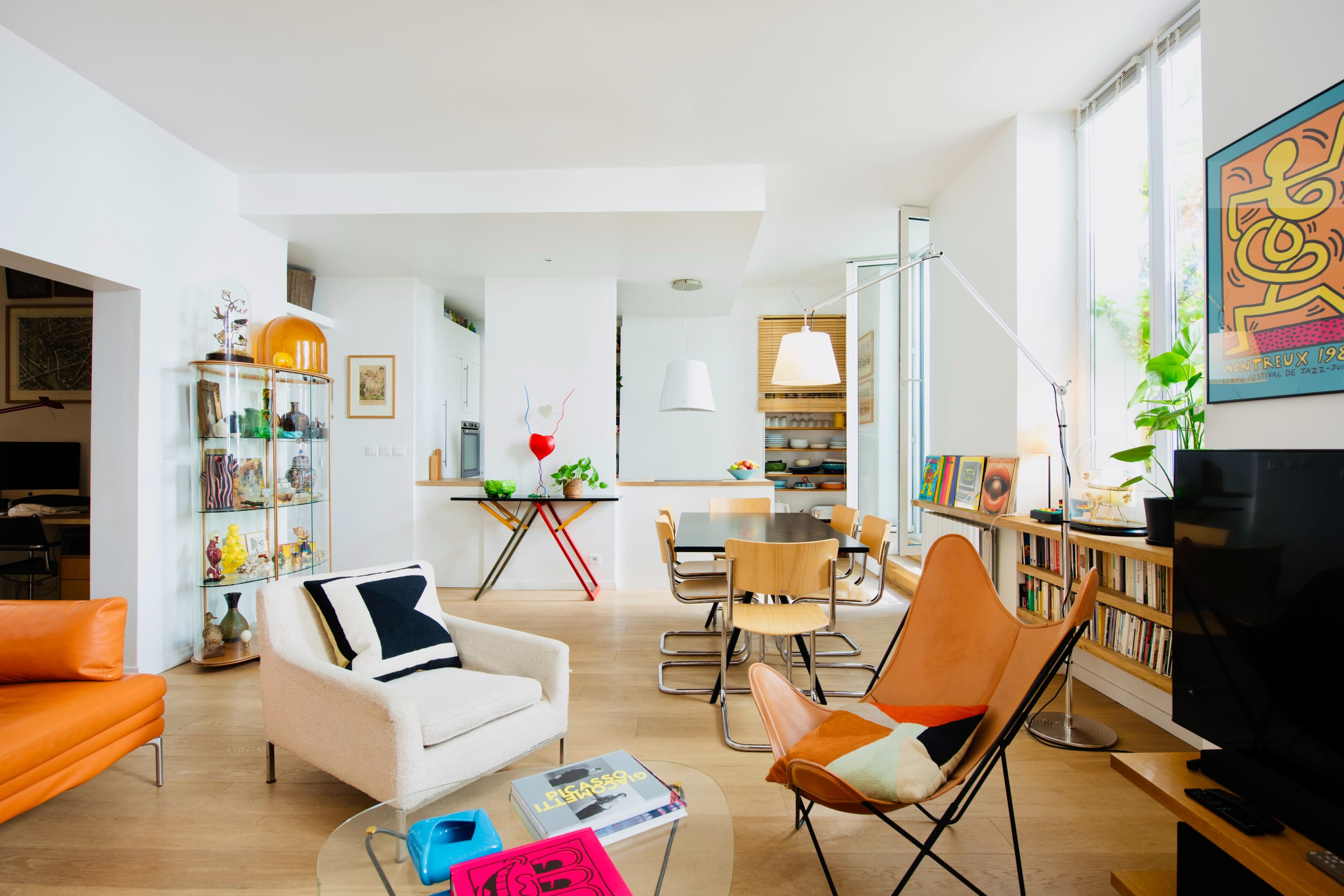 A modern living space features a mix of furniture, including an orange sofa, a white armchair, and wooden dining chairs, alongside shelves filled with books and decorative items.