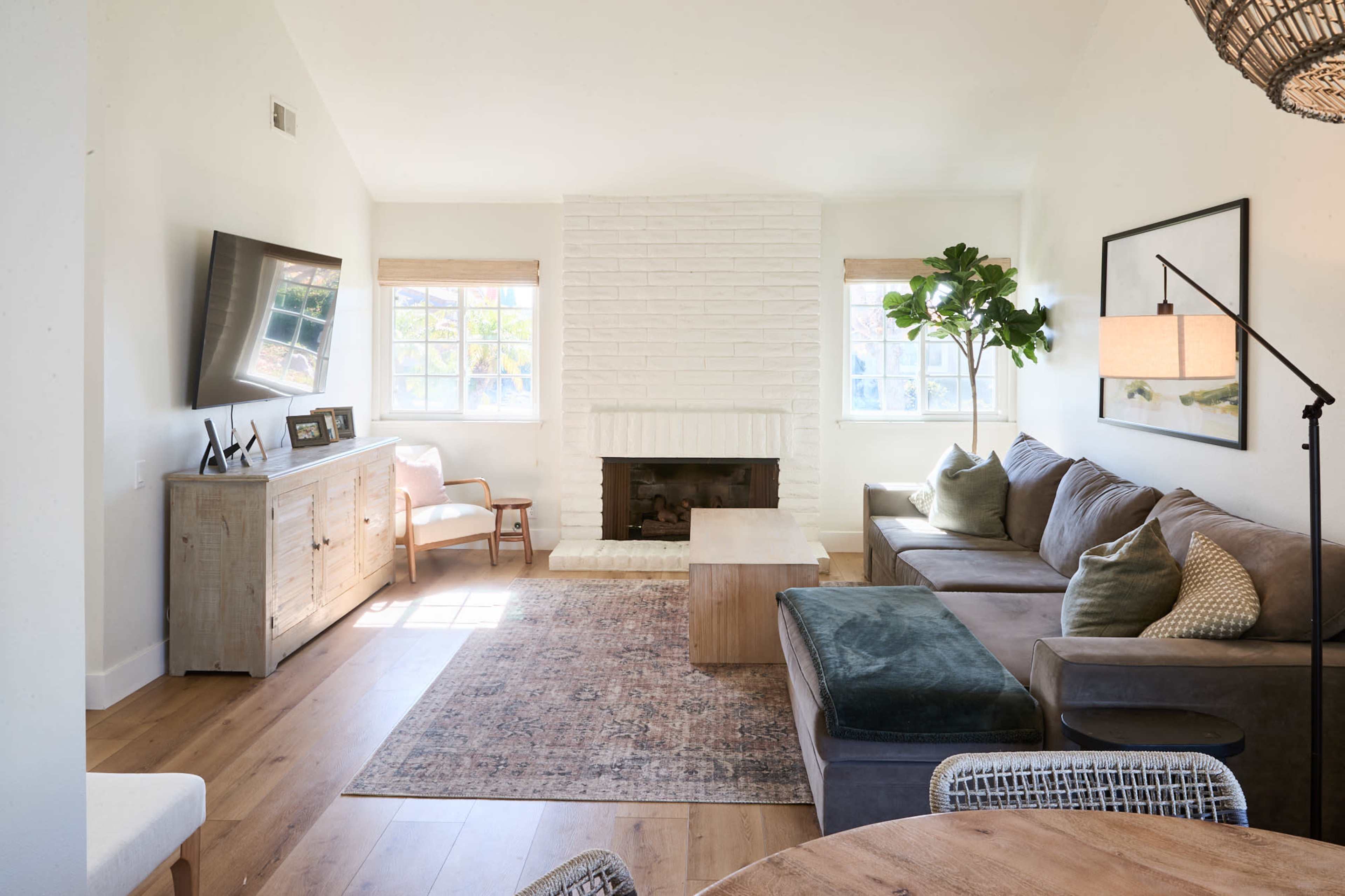 A bright living room featuring a sectional sofa, a wooden coffee table, a white brick fireplace, and large windows with natural light.