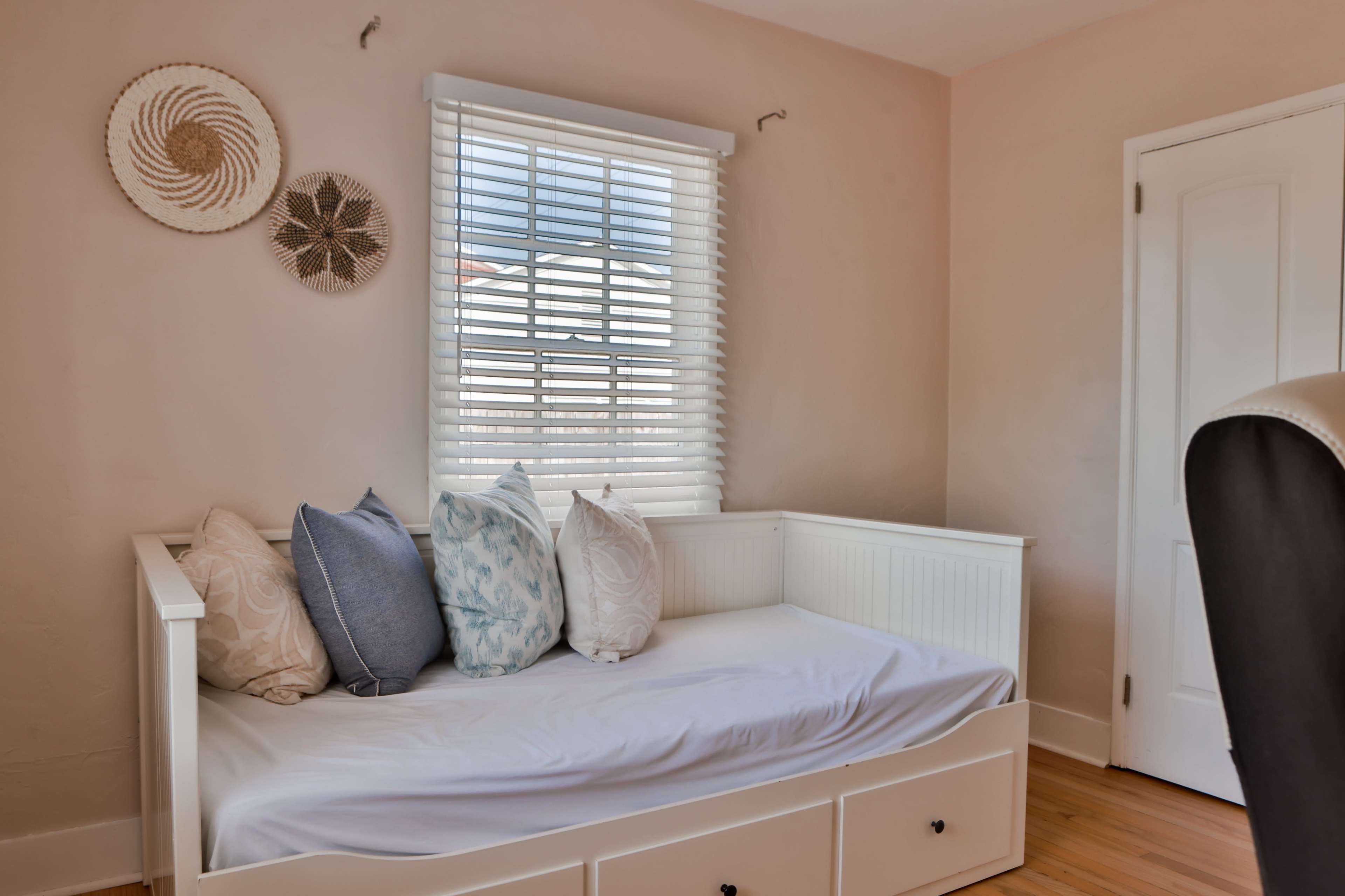 A light-colored room with a daybed adorned with pillows and a window featuring white blinds.