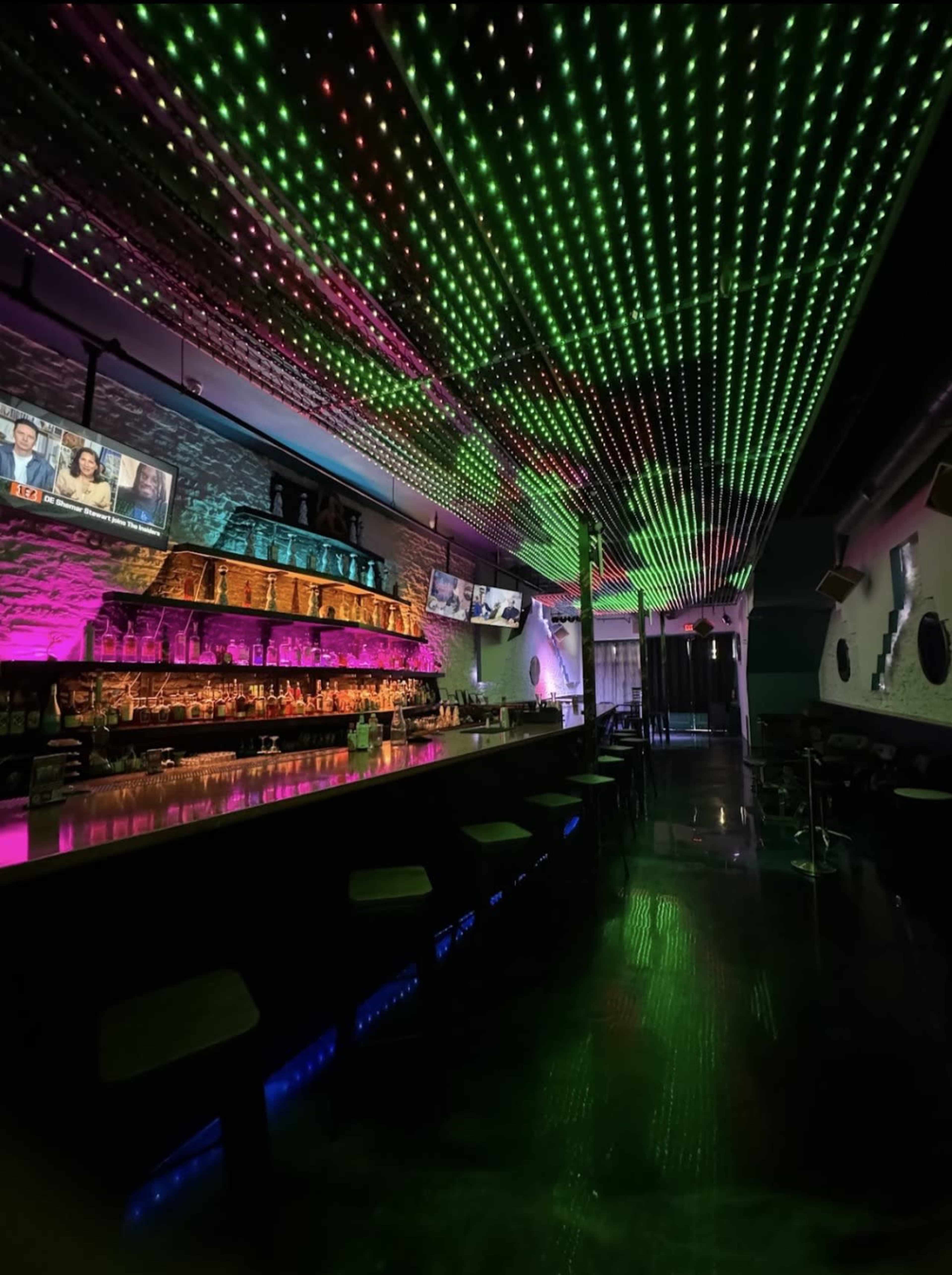 The image shows a modern bar with a colorful LED ceiling, illuminated shelves behind the bar, and multiple screens displaying content.
