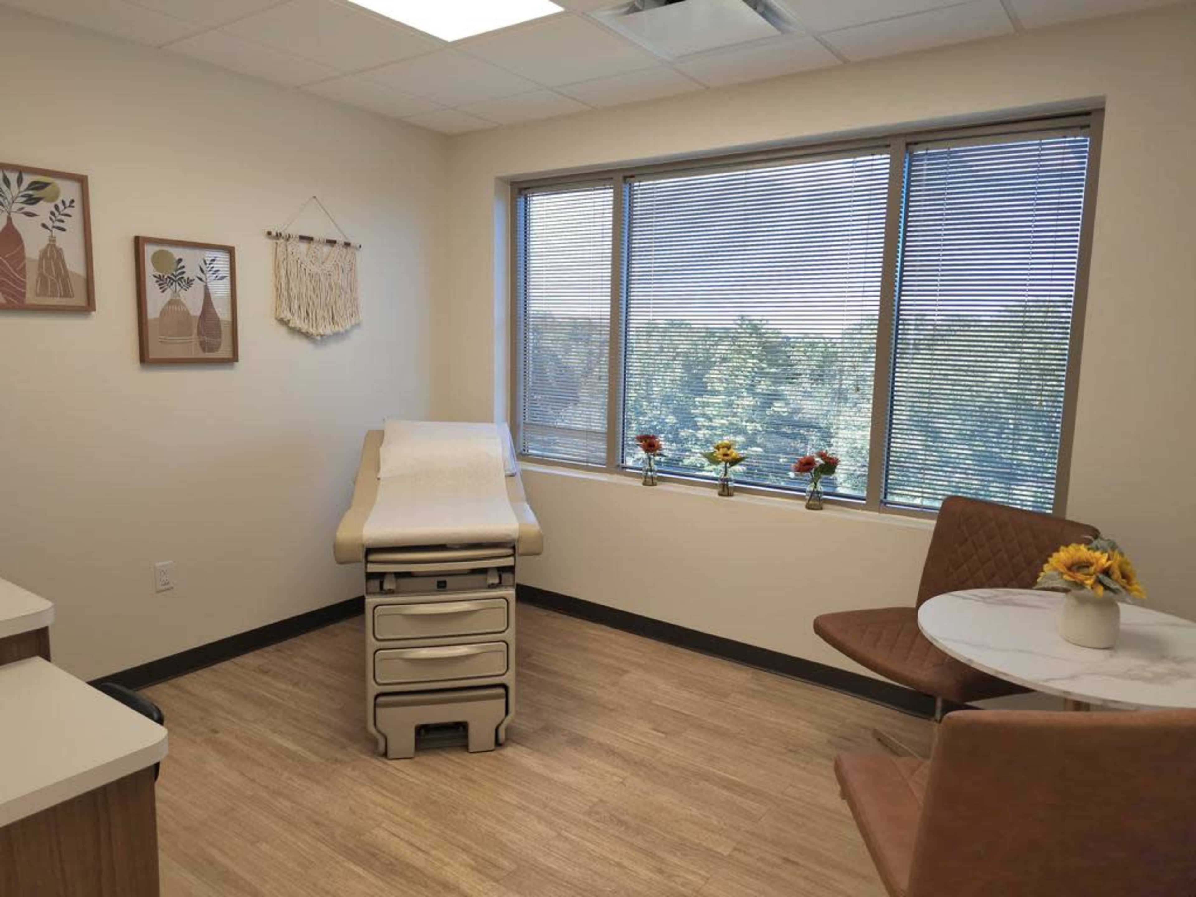 The image shows a medical examination room featuring an examination table, a small round table with two chairs, and a window with a view of greenery outside.