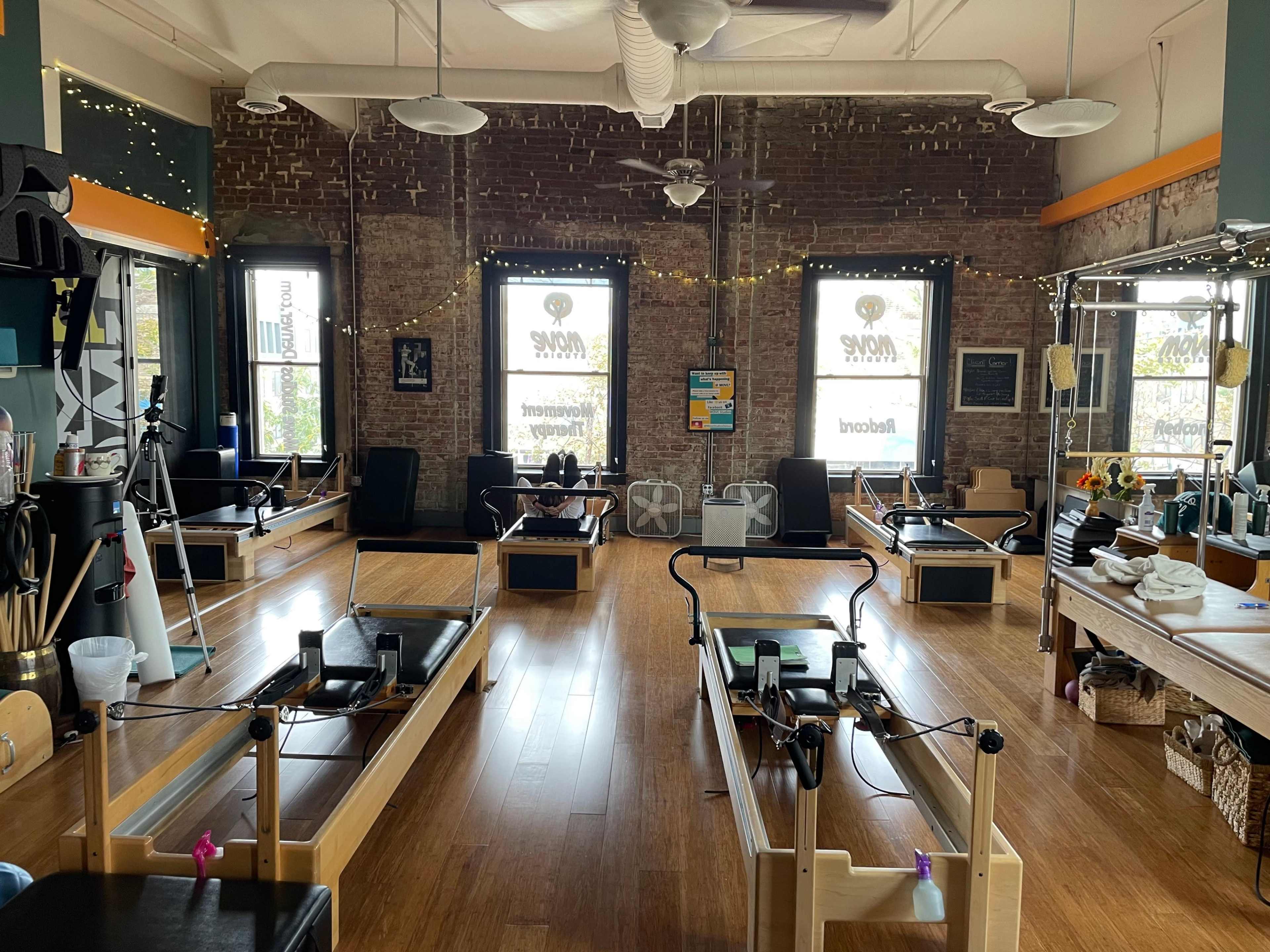 A spacious Pilates studio with wooden floors, several reformer machines, and large windows letting in natural light.