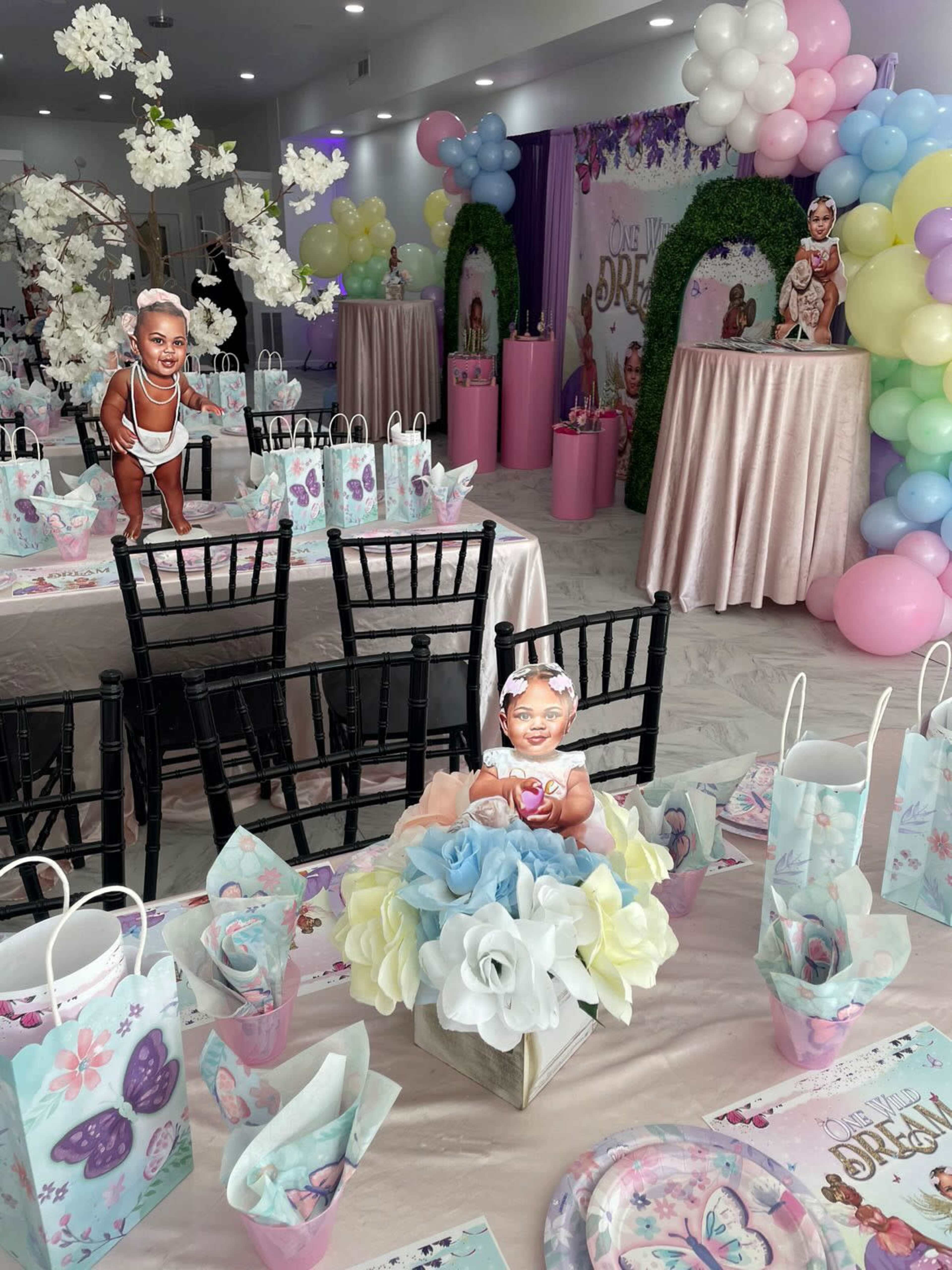 The scene depicts a decorated party venue with tables set for a celebration, featuring pastel colored balloons, floral arrangements, and themed tableware.