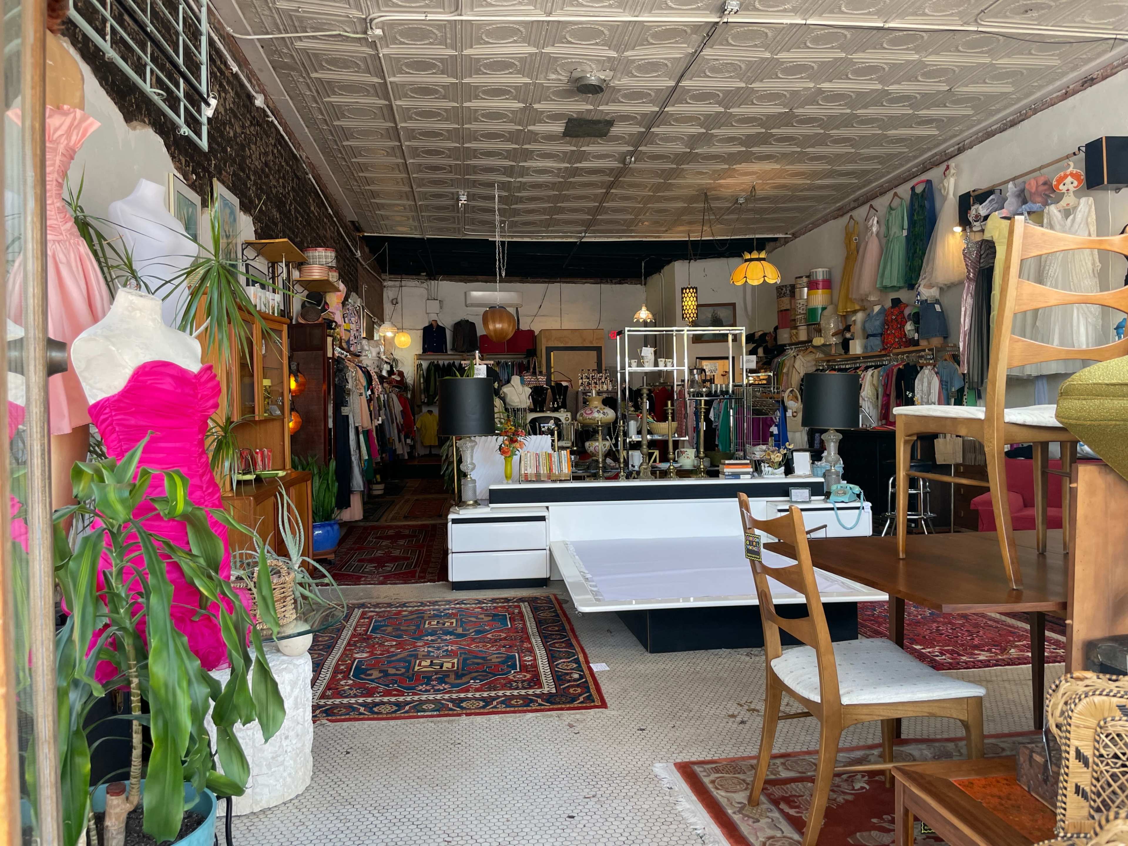 A vintage clothing store displays a variety of garments and accessories on racks, with a mix of furniture and decor items arranged throughout the space.